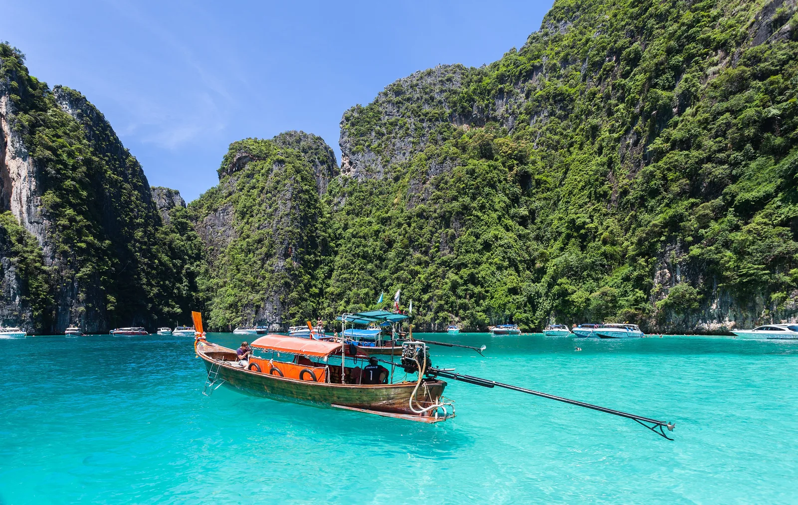 Ein Langheckboot mit orangefarbenem Dach auf türkisfarbenem Wasser vor den bewachsenen Kalkwänden von Ko Phi Phi Le