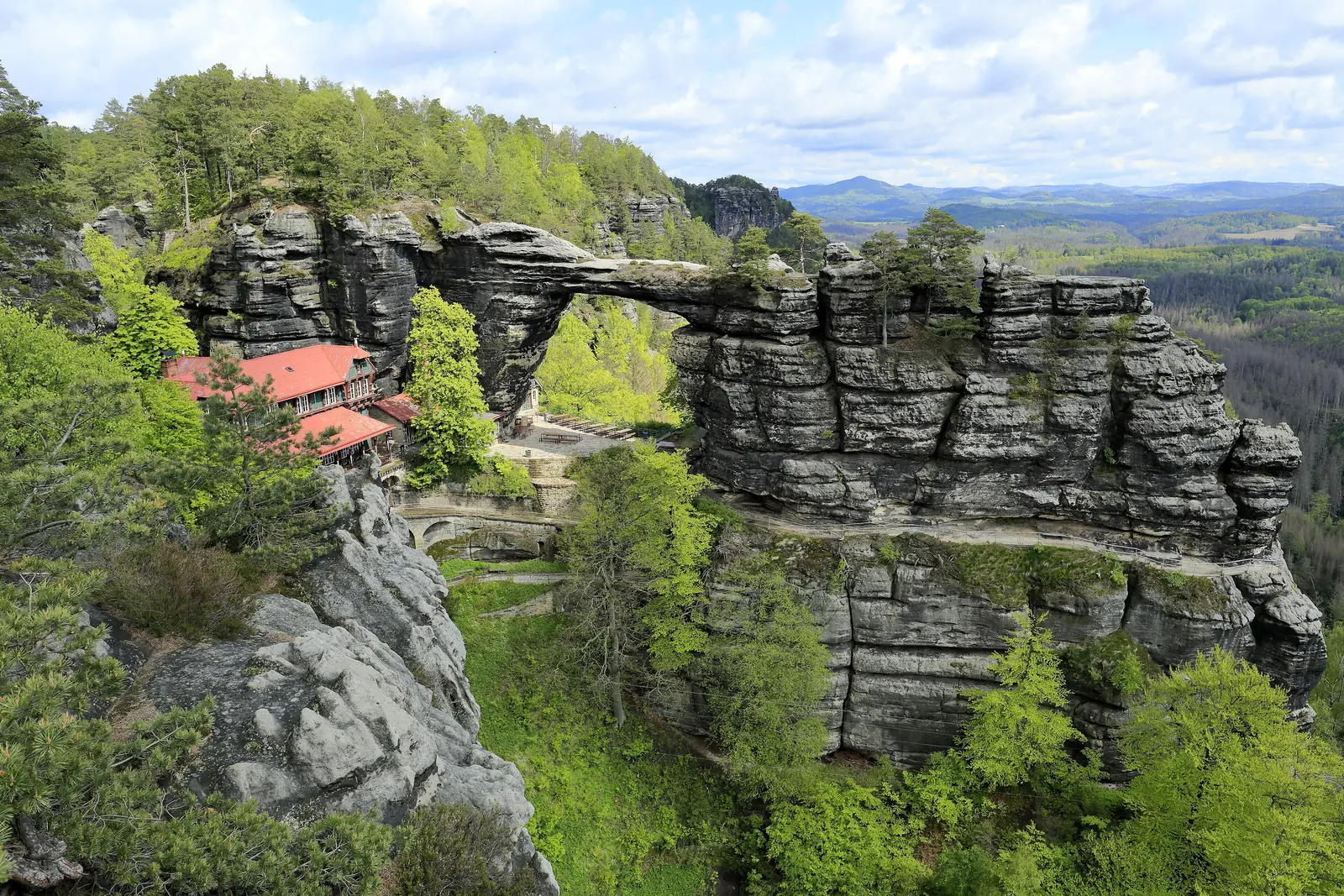 Das Prebischtor vom Aussichtspunkt Kreuzstein: der Sandsteinbogen über dem Falkennest mit roten Dächern, ringsum Wald und Felsen