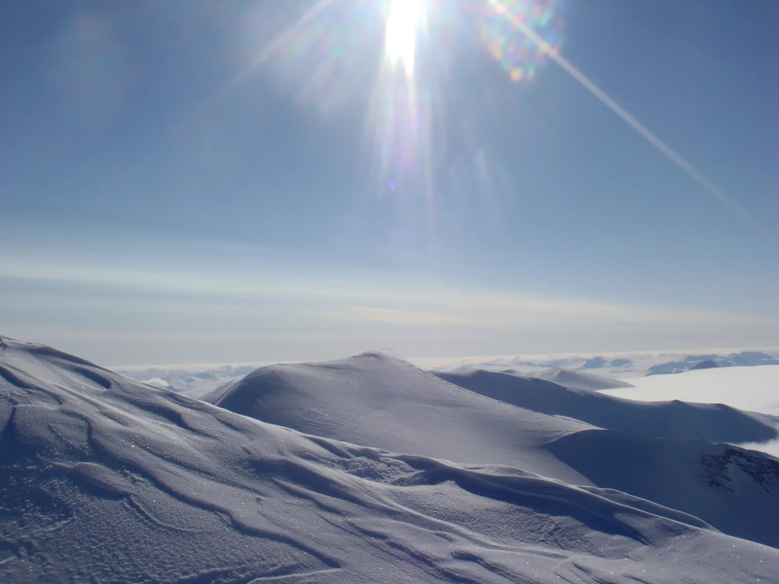 Blick vom Gipfel über verwehte Schneerücken auf ein weites Eisfeld, darüber die tief stehende Sonne am blauen Himmel