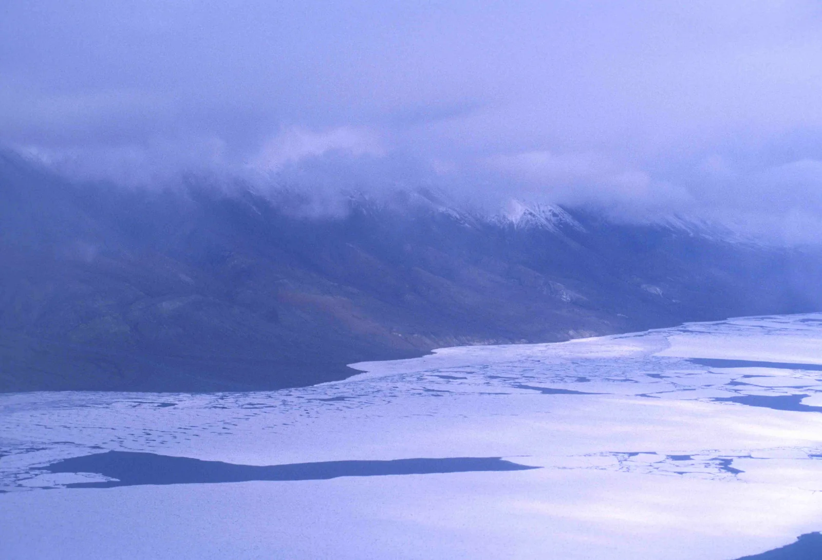 Der zugefrorene Lake Hazen mit aufgebrochenen Eisschollen, dahinter eine langgezogene Bergkette unter tief hängenden Wolken