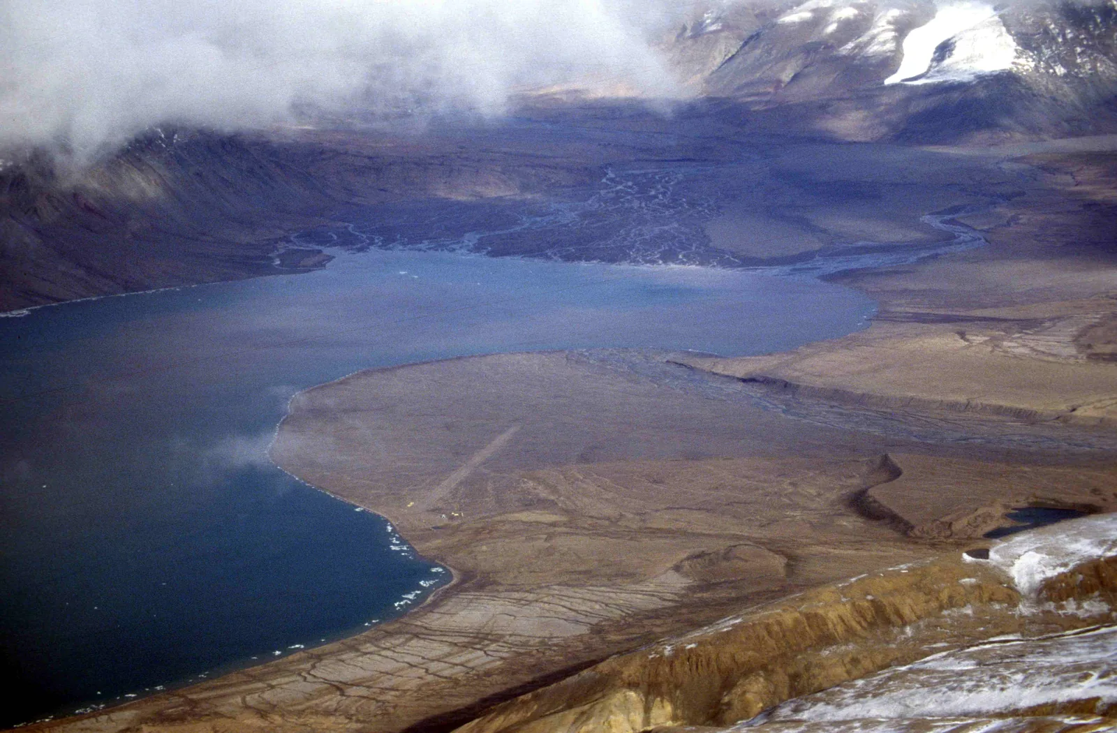 Luftbild auf das Ende eines Fjords: dunkles Wasser, davor eine flache Schotterebene mit Landepiste, ringsum kahle Berge