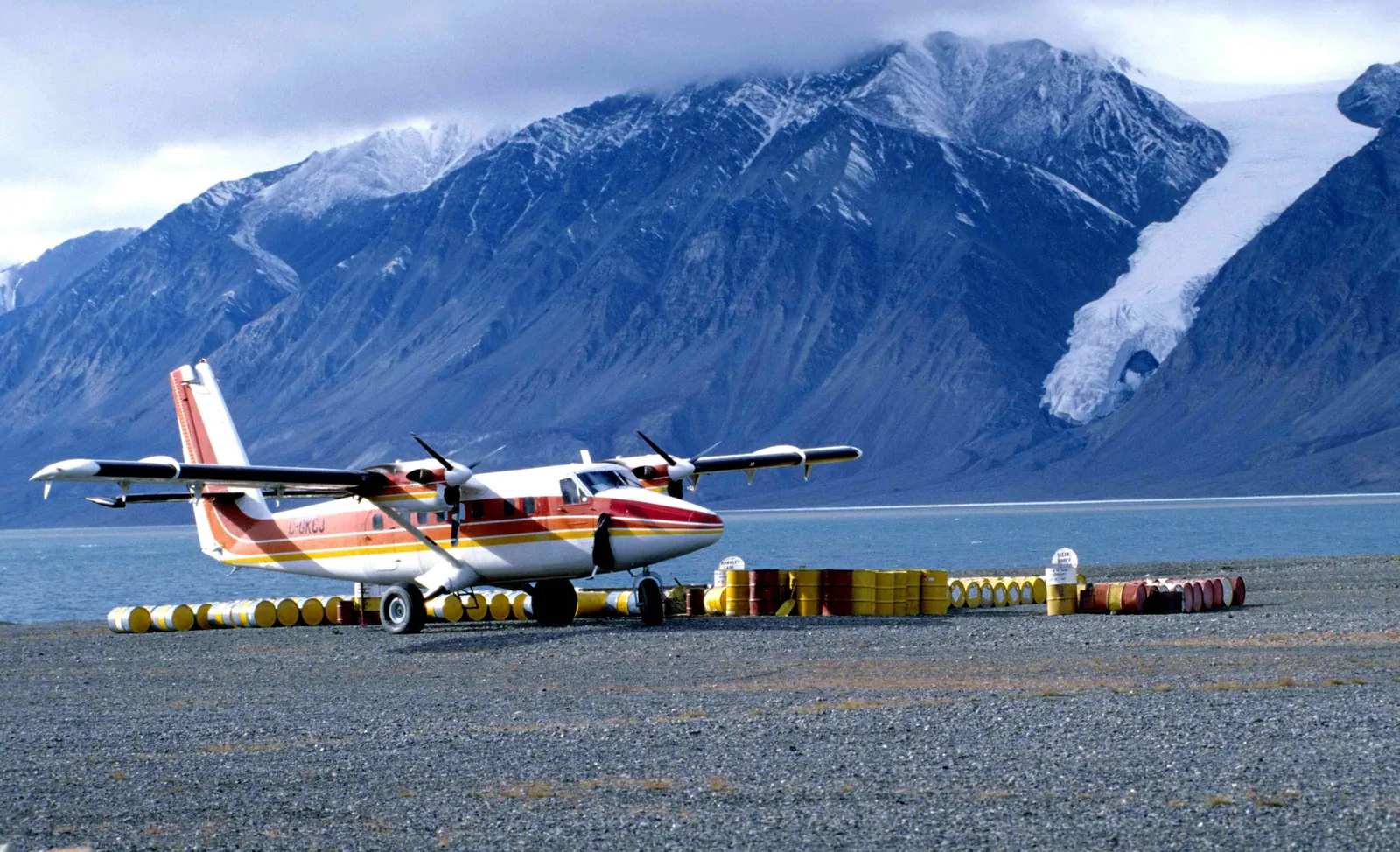 Eine rot-weiße Twin Otter steht auf einer Schotterpiste zwischen Fassreihen, dahinter ein Fjord und vergletscherte Berge