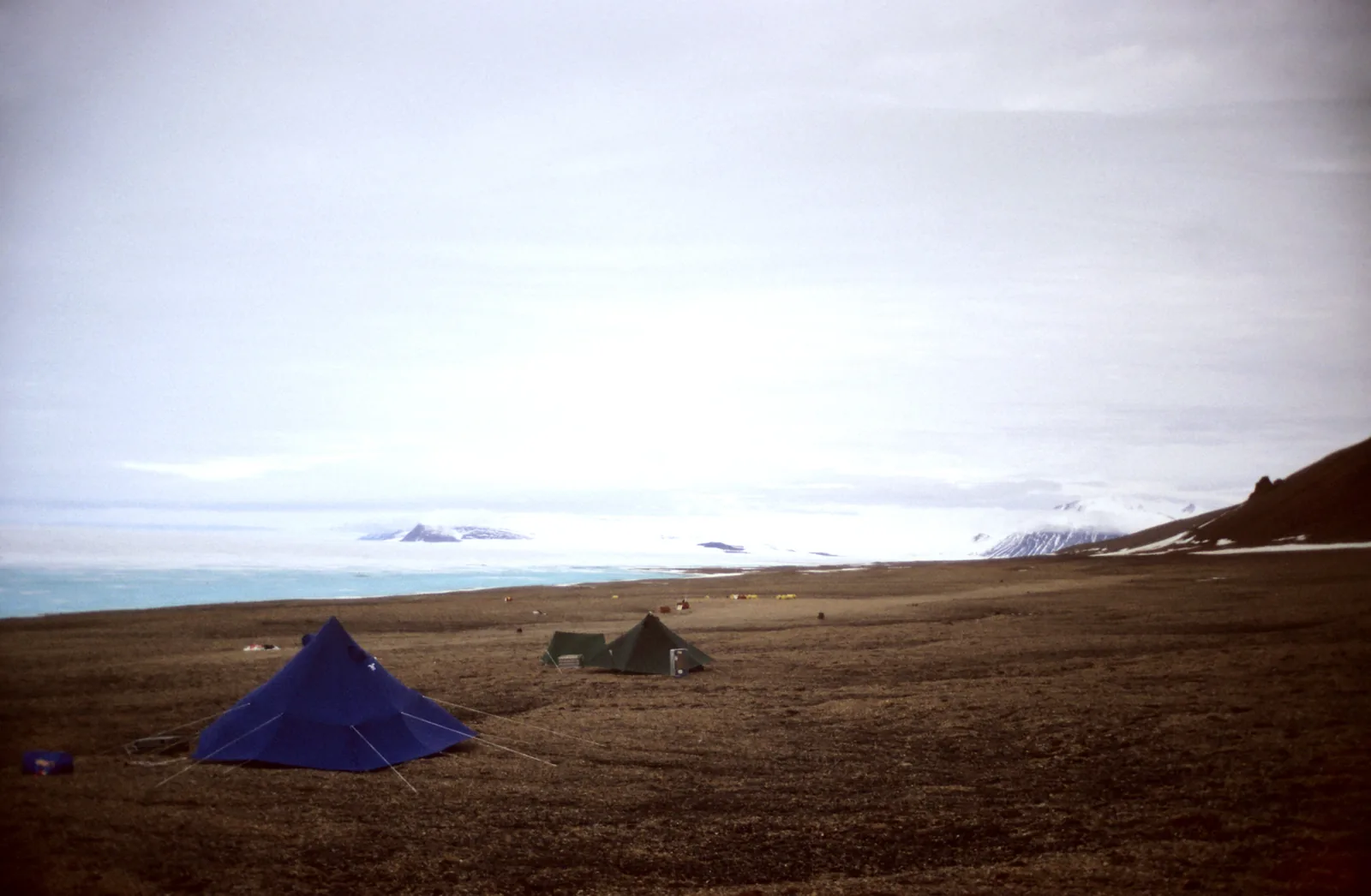 Zwei Zelte eines Feldlagers auf brauner Tundra, dahinter die weiße Fläche des Schelfeises und ferne Berge