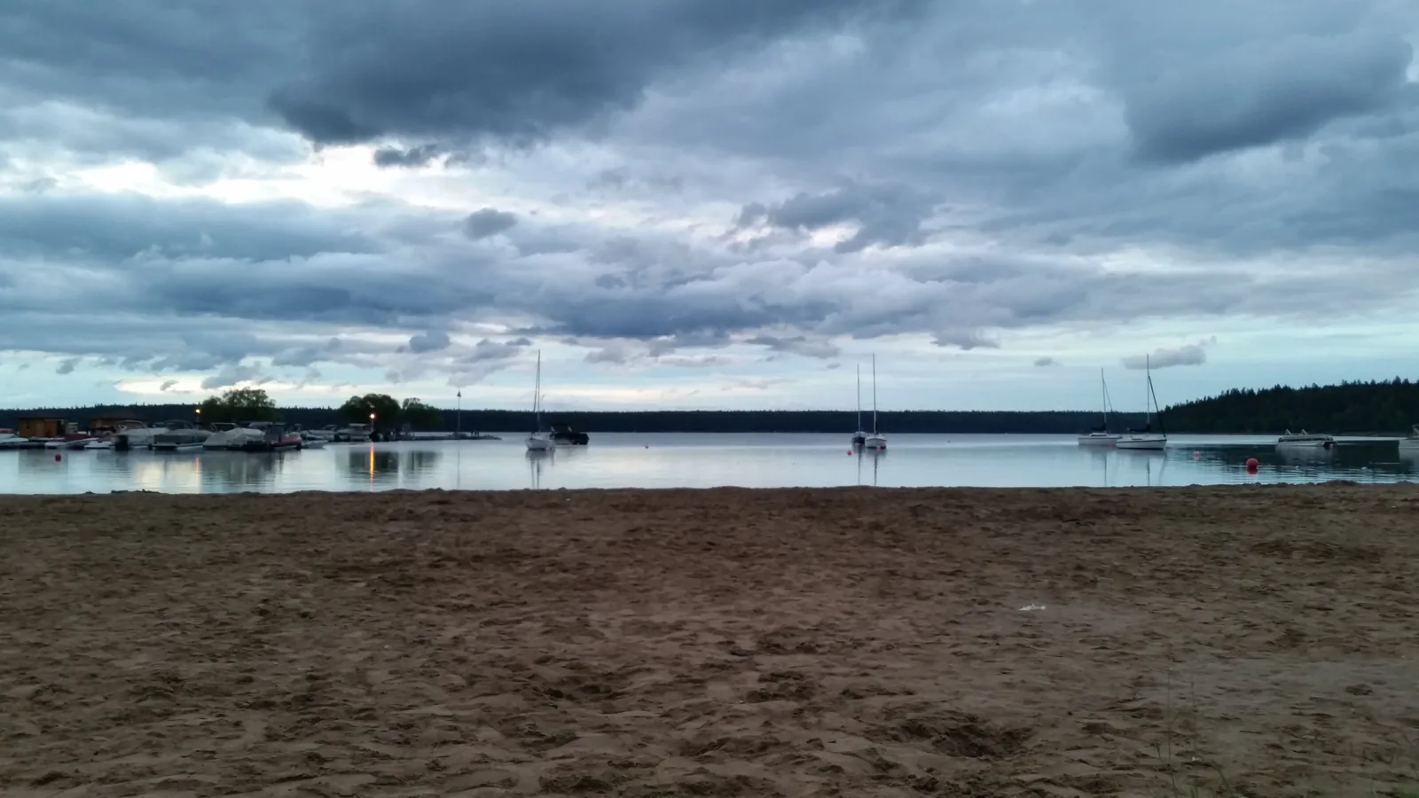 Der Hauptstrand von Wasagaming unter einem schweren Wolkenhimmel, am Wasser liegen Segelboote, der Sand ist leer