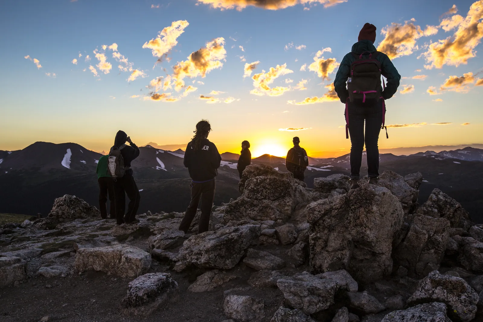 Mehrere Menschen stehen als Silhouetten auf Felsblöcken und blicken über die Tundra auf einen orangefarbenen Sonnenuntergang hinter Bergketten