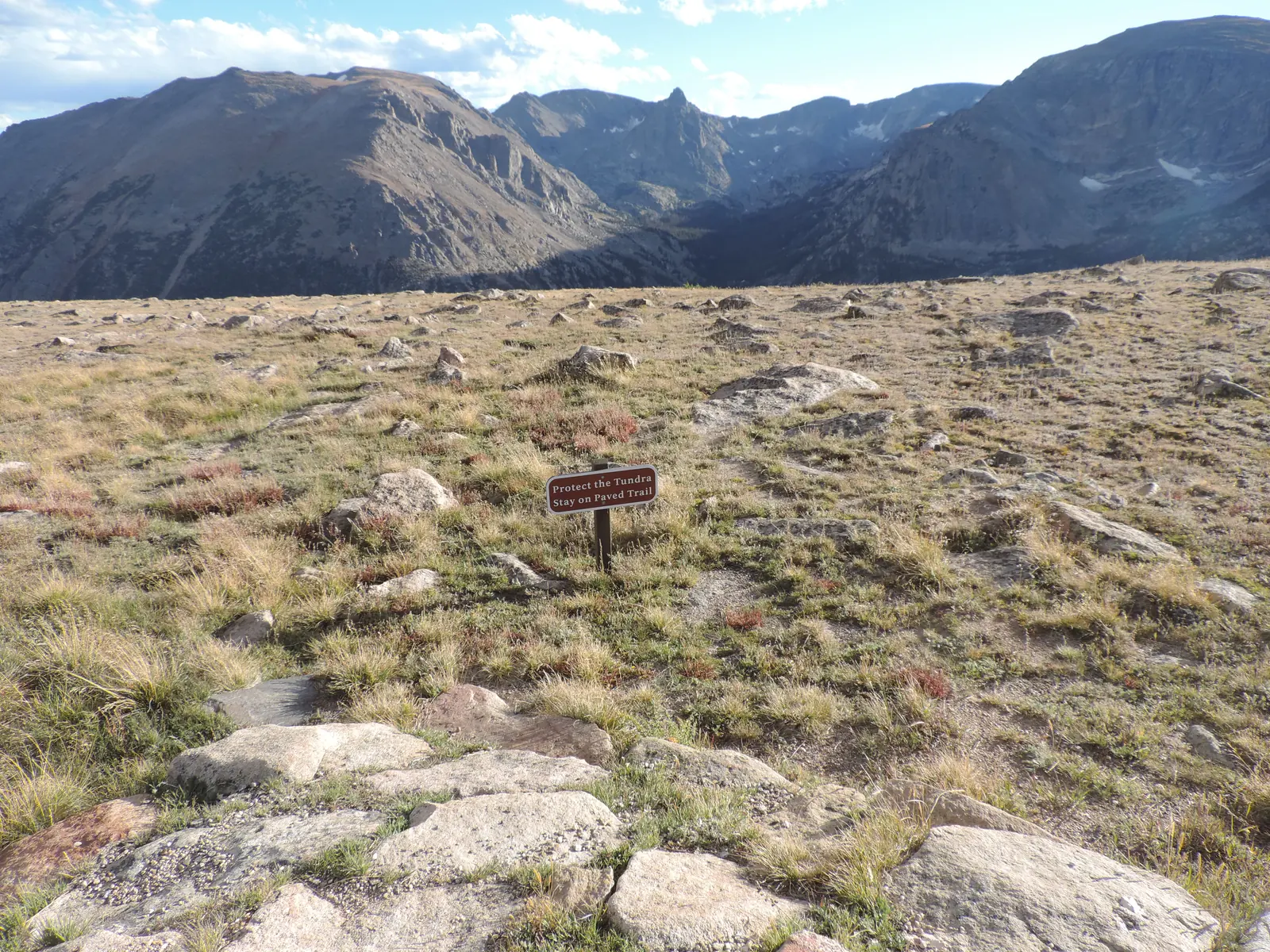 Karge Hochfläche mit niedrigem Gras und verstreuten Granitblöcken, in der Mitte ein kleines braunes Schild, im Hintergrund kahle Bergkämme