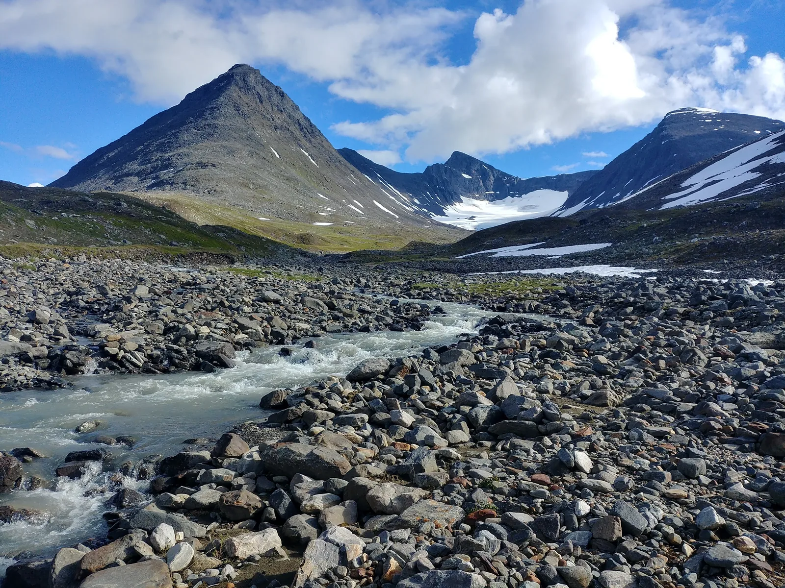 Ein Gletscherbach fließt durch ein Geröllfeld im Tal Bastavagge im Sarek-Nationalpark, dahinter ein spitzer Berg und Schneefelder