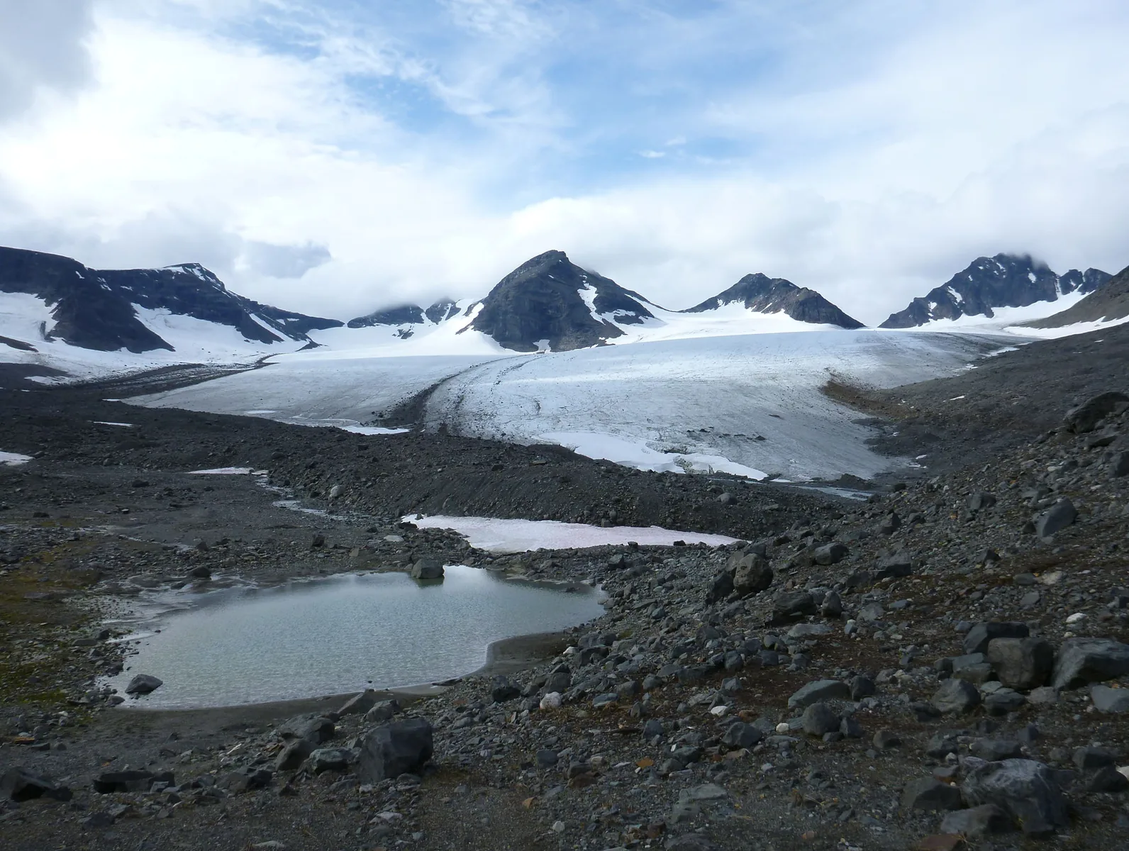 Die breite Gletscherzunge des Bårddejiegŋa im Sarek-Nationalpark, davor Moränenschutt und kleine Schmelzwasserseen, dahinter dunkle Gipfel