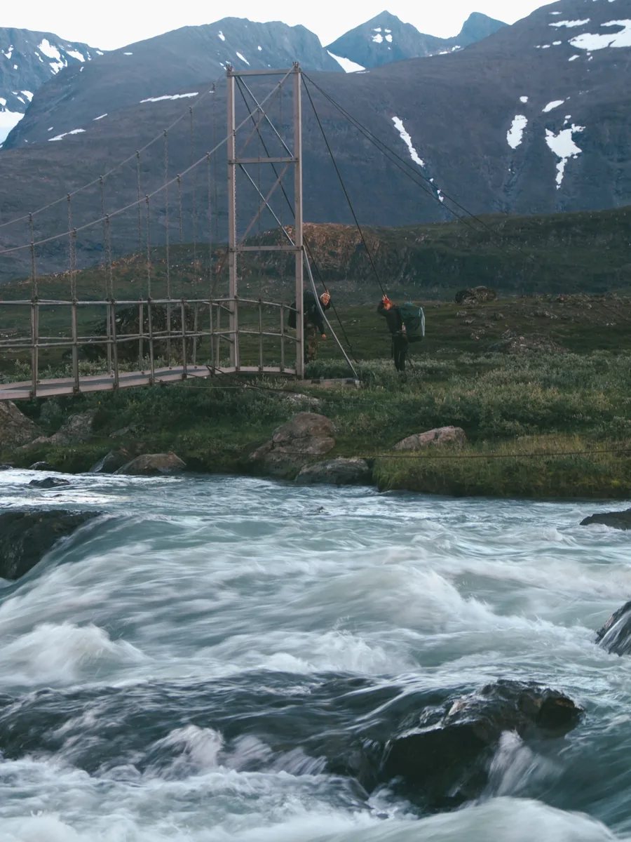 Eine schmale Hängebrücke führt im Sarek-Nationalpark über einen tosenden Gebirgsbach, dahinter Wanderer und schneefleckige Berge