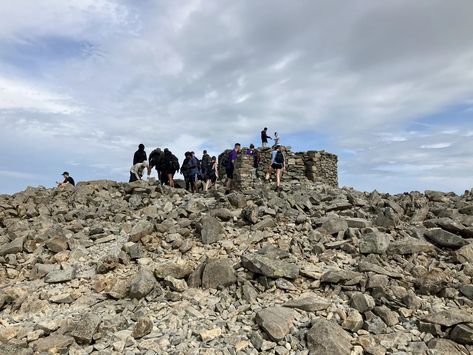 Eine Wandergruppe am runden Steinmann auf dem Gipfel des Scafell Pike im Lake-District-Nationalpark, ringsum Geröll