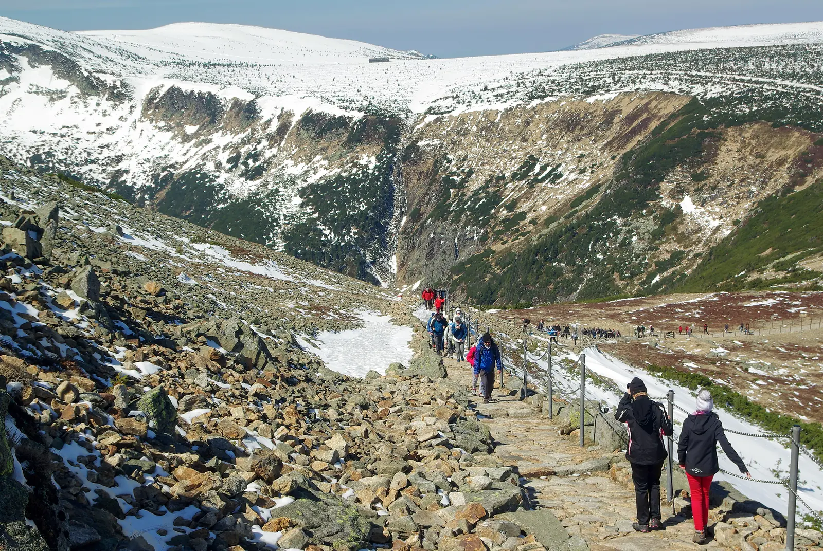 Wanderer auf dem gepflasterten Weg zur Schneekoppe zwischen Schneeresten, links der Abbruch in den Kessel