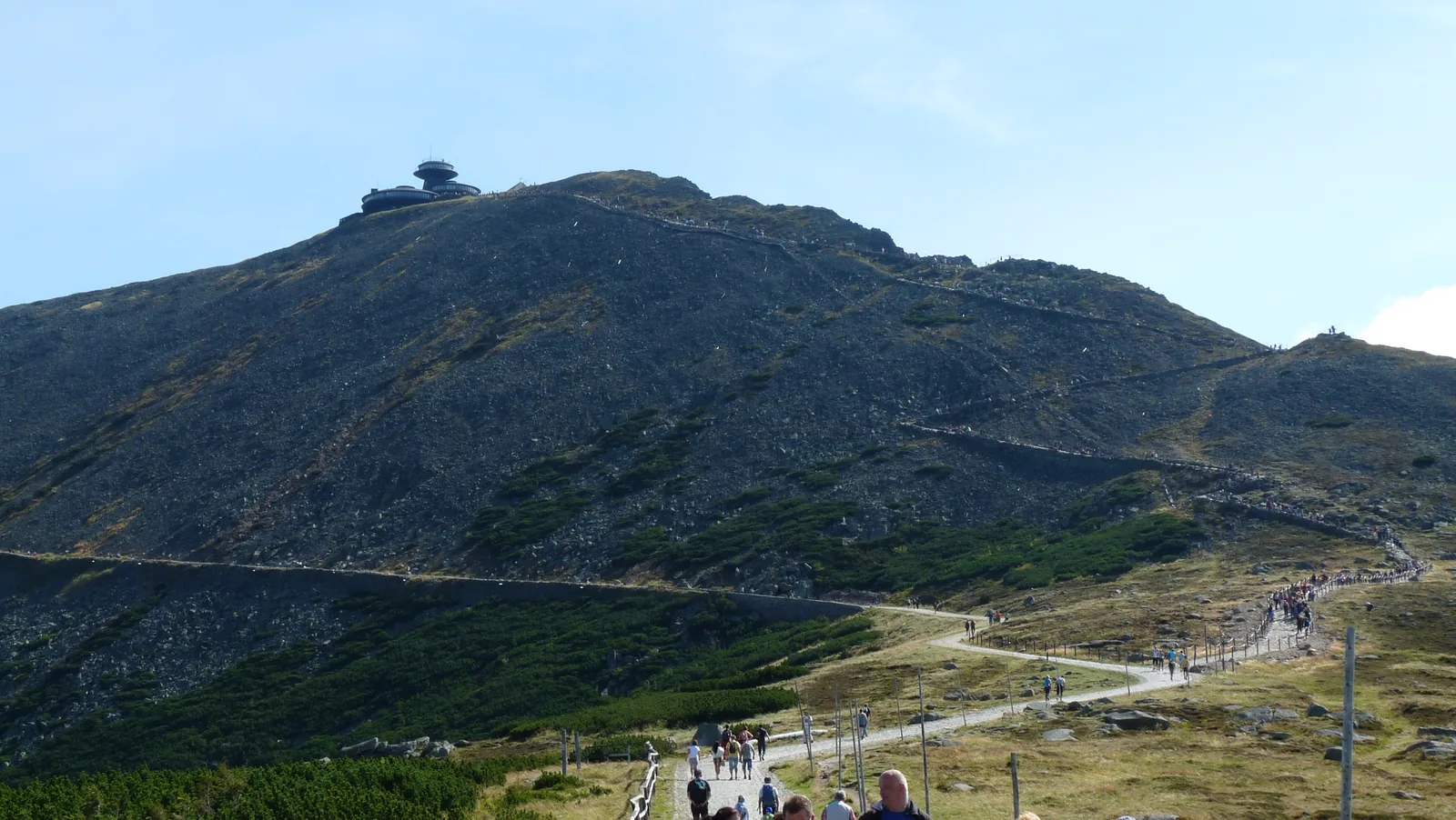 Der Gipfelkegel der Schneekoppe im Riesengebirge mit den Gebäuden oben, davor der Weg mit Wanderern auf dem Kamm