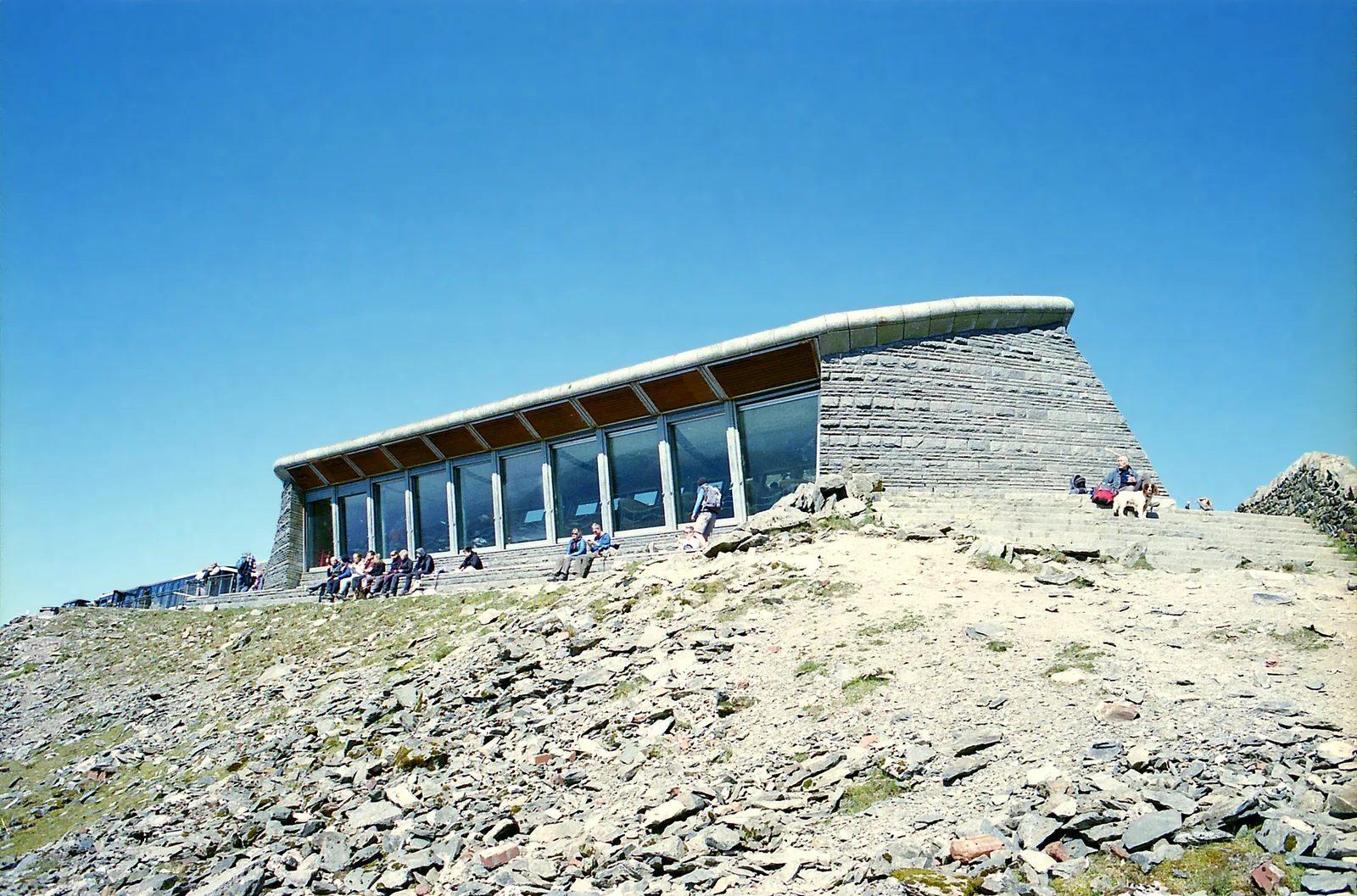 Das Gipfelgebäude Hafod Eryri auf dem Snowdon im Snowdonia-Nationalpark mit großer Fensterfront, davor Wanderer