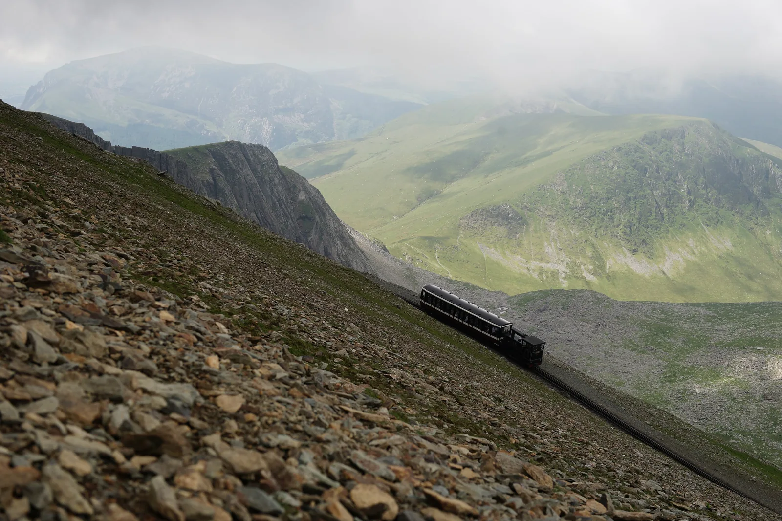 Ein Zug der Snowdon Mountain Railway am steinigen Hang des Snowdon im Snowdonia-Nationalpark, dahinter Täler und Wolken