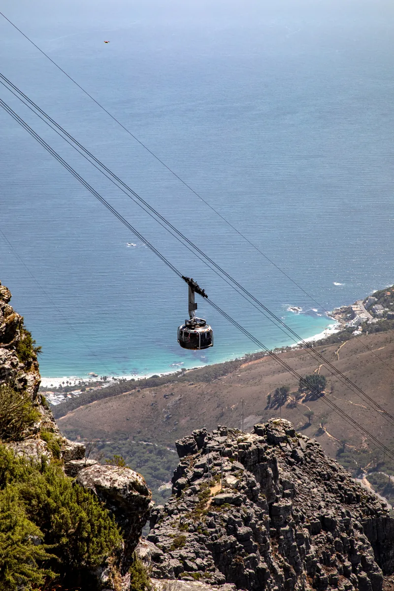 Eine runde Gondel der Tafelberg-Seilbahn hängt an zwei Seilen über einer Felskante, tief darunter Küste und Meer bei Camps Bay