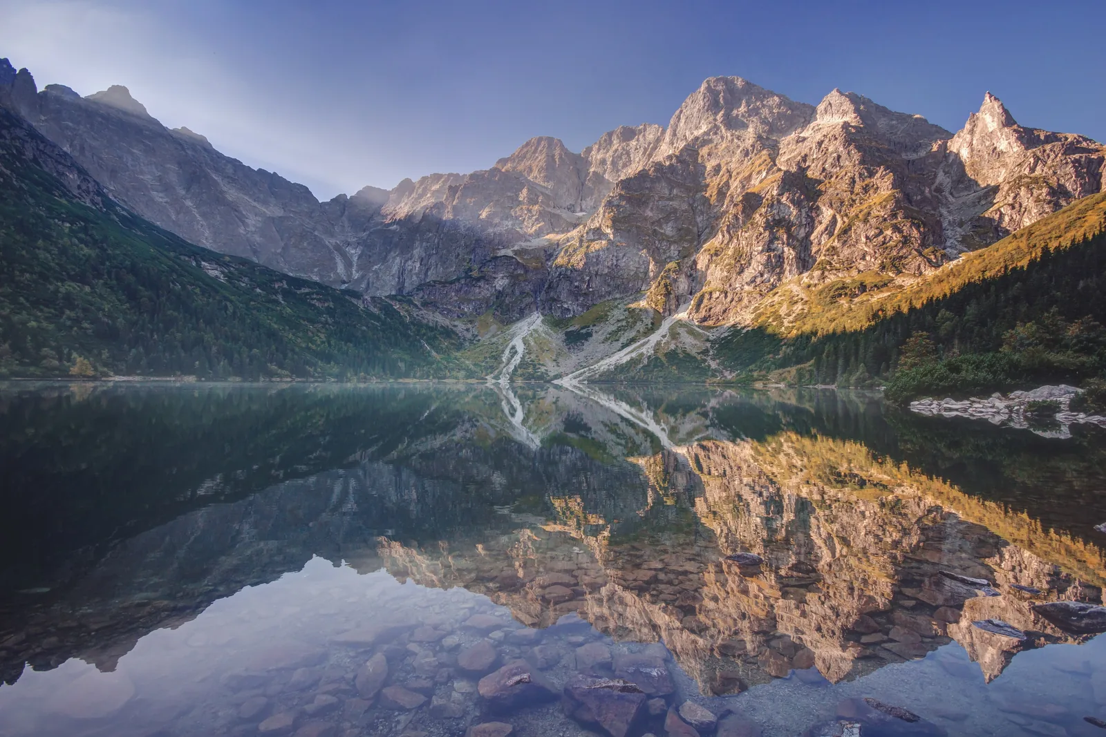 Der Morskie Oko im Tatra-Nationalpark am Morgen: Die Gipfel über dem See spiegeln sich im glatten Wasser, rechts bewaldete Hänge