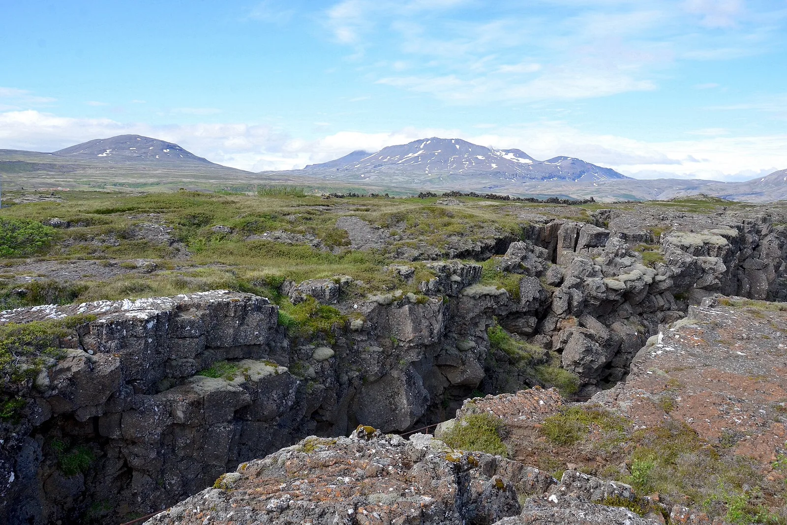 Die Almannagjá im Nationalpark Þingvellir: eine von Moos bewachsene Felsspalte im Lavafeld, dahinter Berge mit Schneeresten
