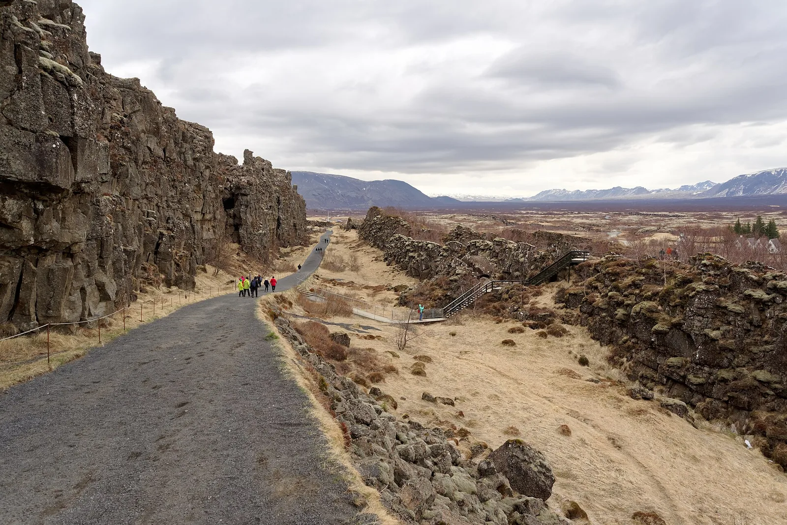 Weg durch die Almannagjá im Nationalpark Þingvellir: links die hohe Basaltwand, rechts der Grabenbruch mit trockenem Gras, Besucher in der Ferne