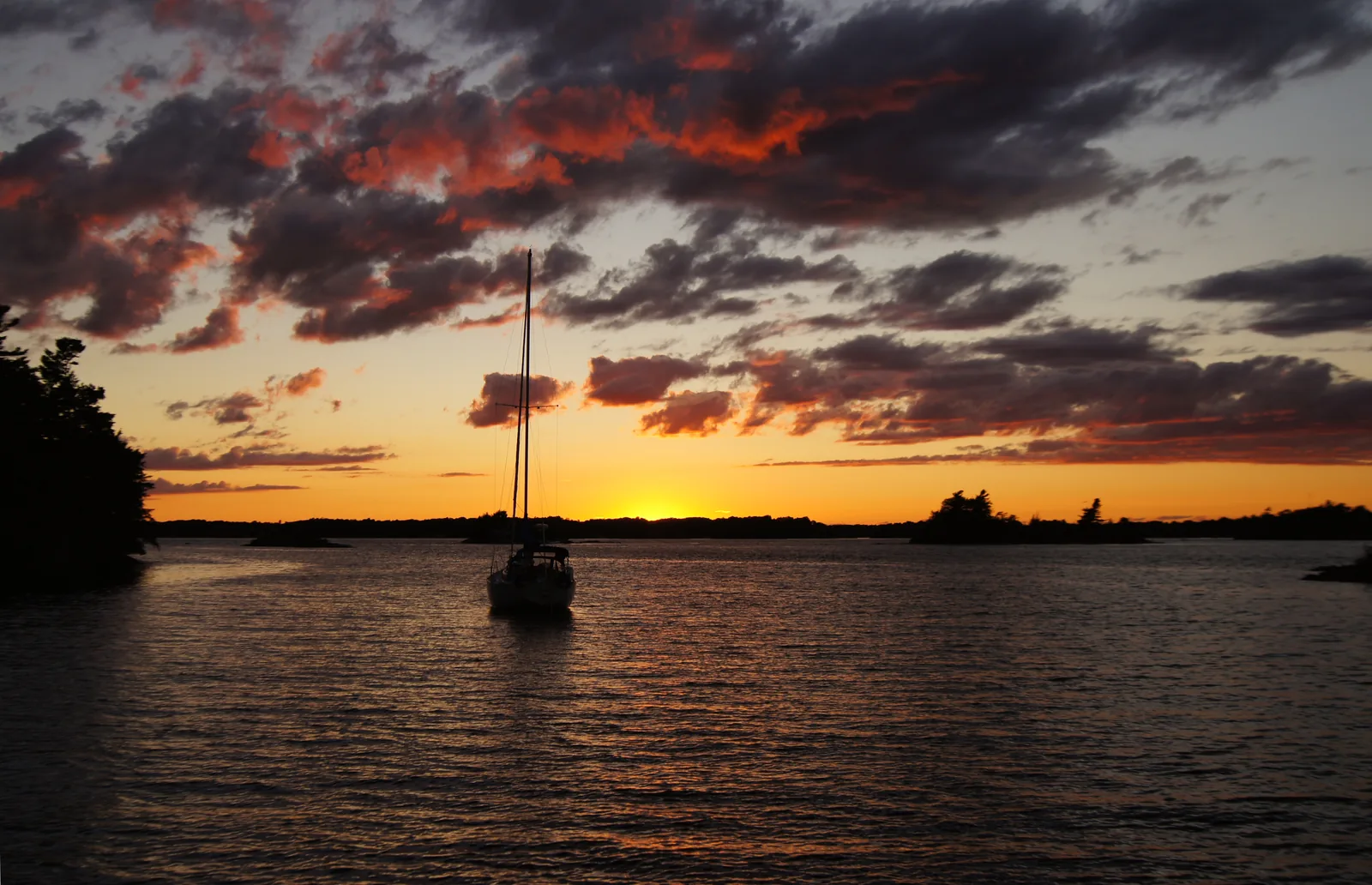 Ein Segelboot liegt bei Sonnenuntergang vor einer bewaldeten Insel, der Himmel ist orange und violett bewölkt
