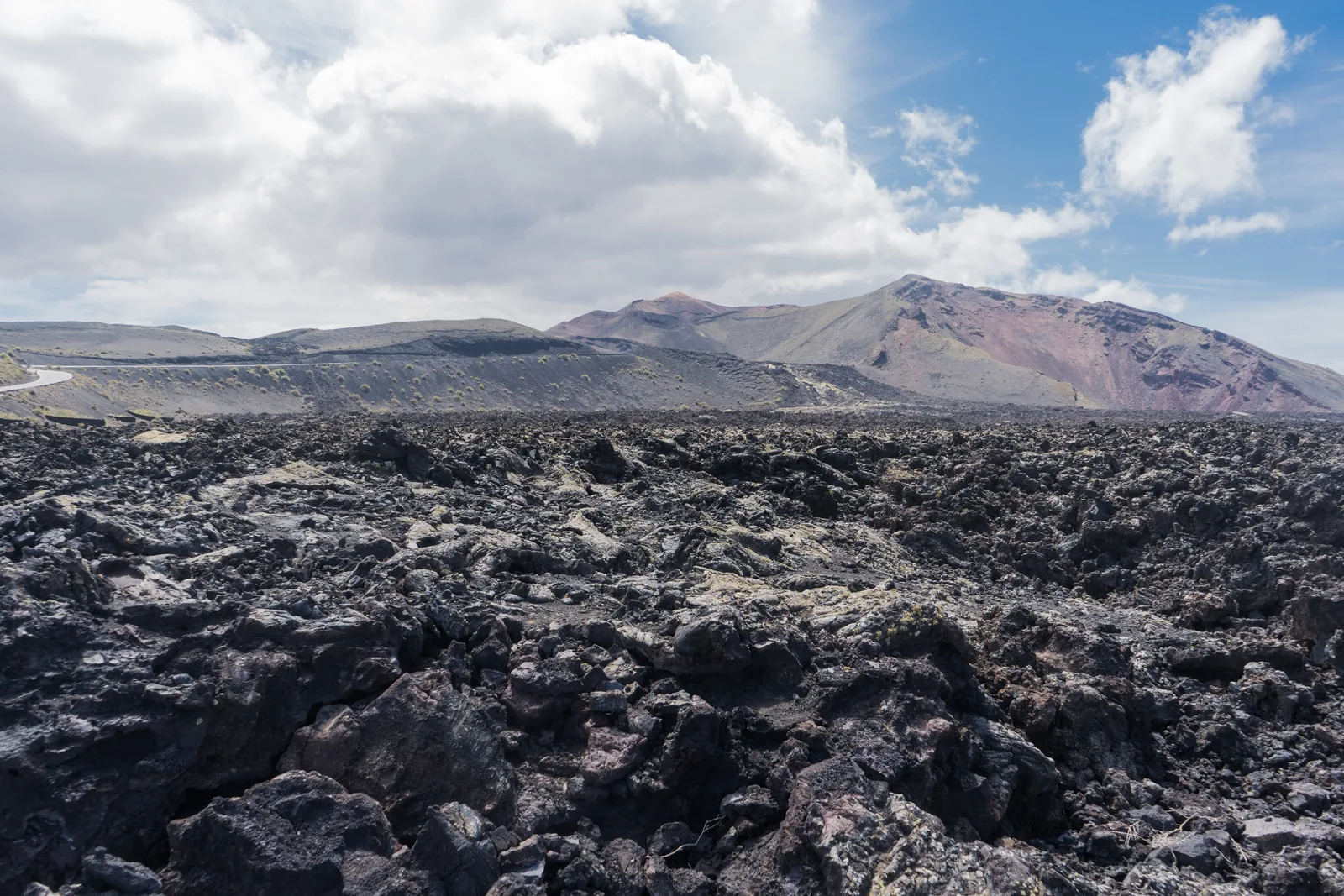Schwarzes Aa-Lavafeld bis zum Horizont, dahinter die bunten Vulkankegel des Timanfaya unter Wolken