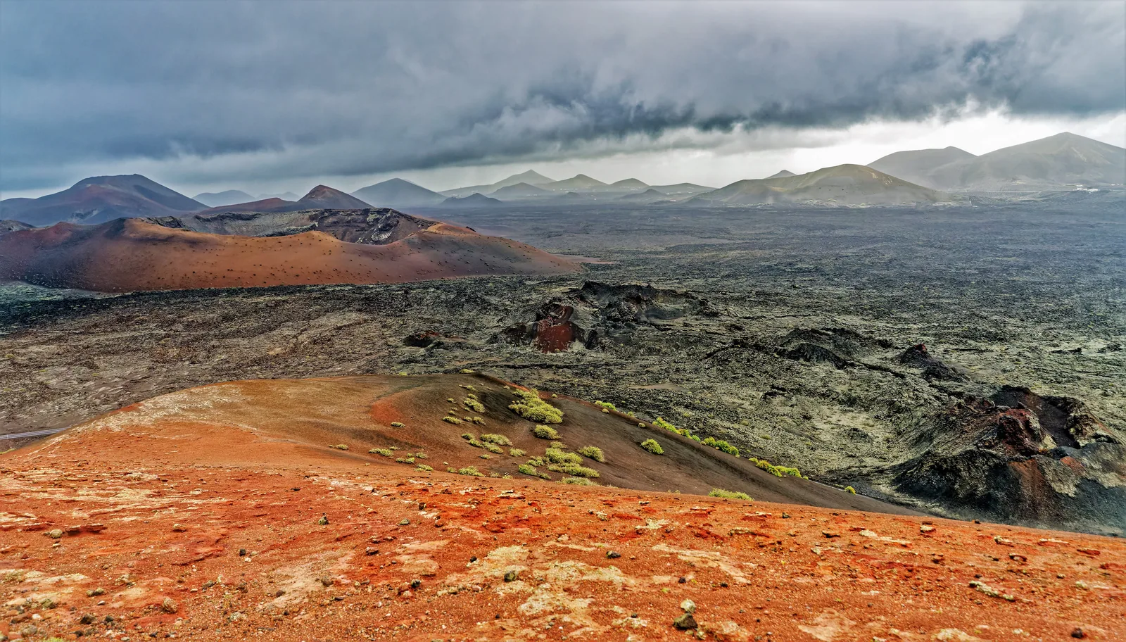 Rote und schwarze Vulkankegel des Timanfaya-Nationalparks unter einer dunklen Wolkendecke, im Vordergrund Lapilli mit ersten Flechten