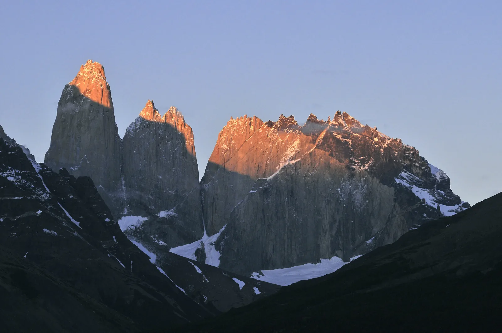 Die Torres del Paine im Nationalpark Torres del Paine bei Sonnenaufgang: die Granittürme und Gipfel glühen orange, darunter dunkle Hänge mit Schneeresten