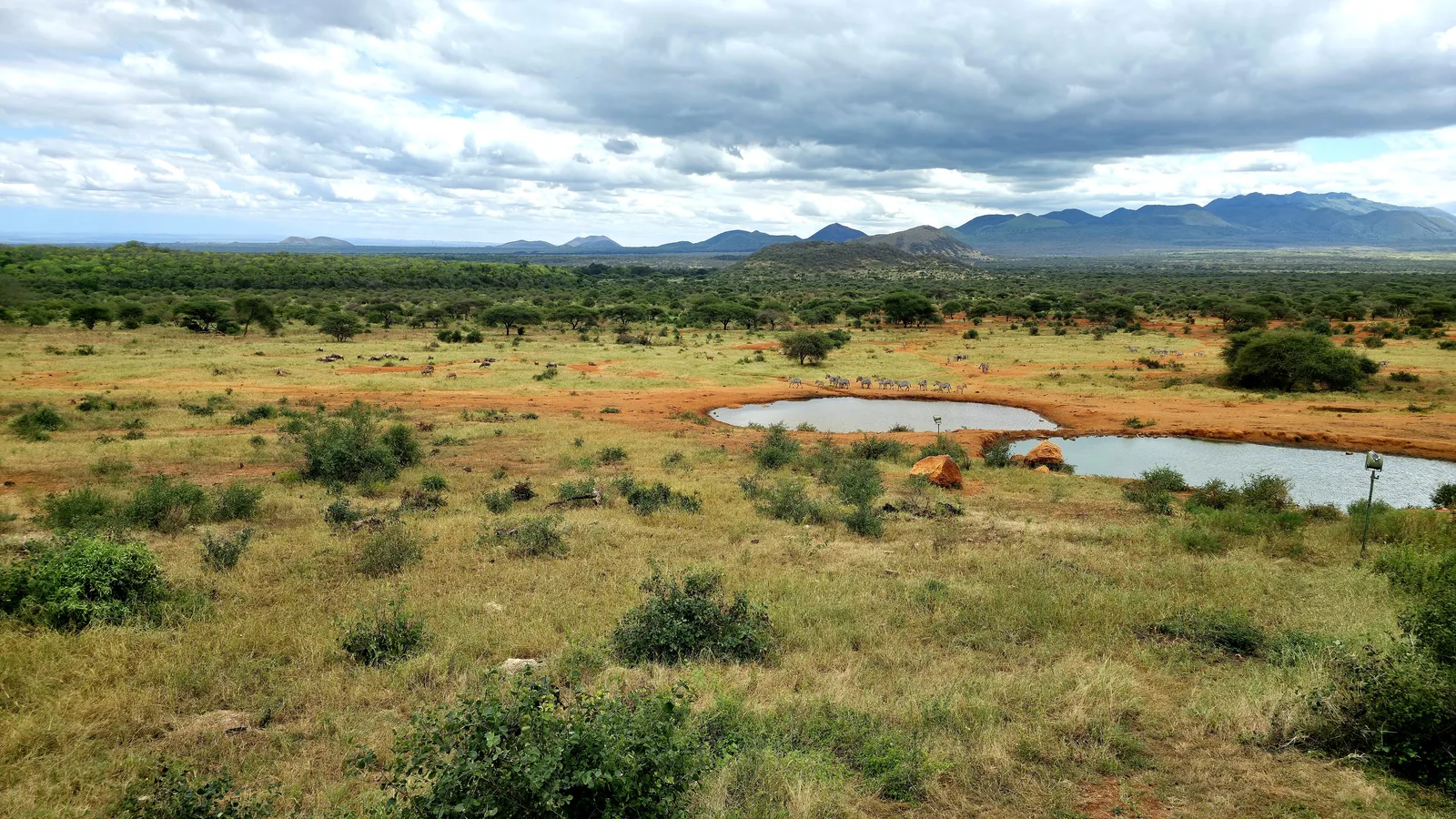 Weite Buschlandschaft im Tsavo-West-Nationalpark mit zwei Wasserlöchern, dahinter Hügel und Berge unter Wolken
