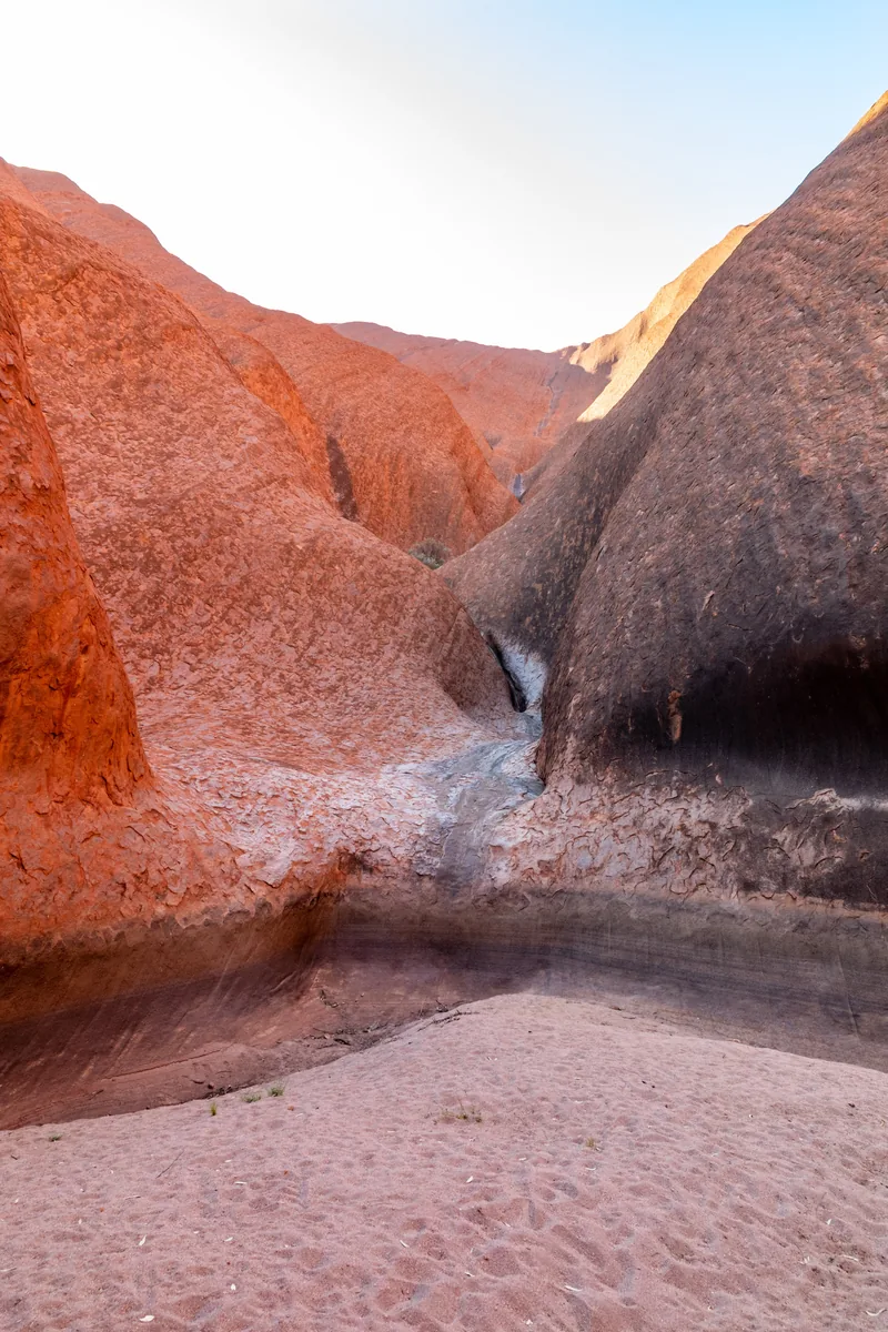 Das Mutitjulu Waterhole am Uluru im Uluṟu-Kata-Tjuṯa-Nationalpark: eine Felsrinne zwischen zwei rötlichen Felswänden mündet in ein Becken, davor heller Sand