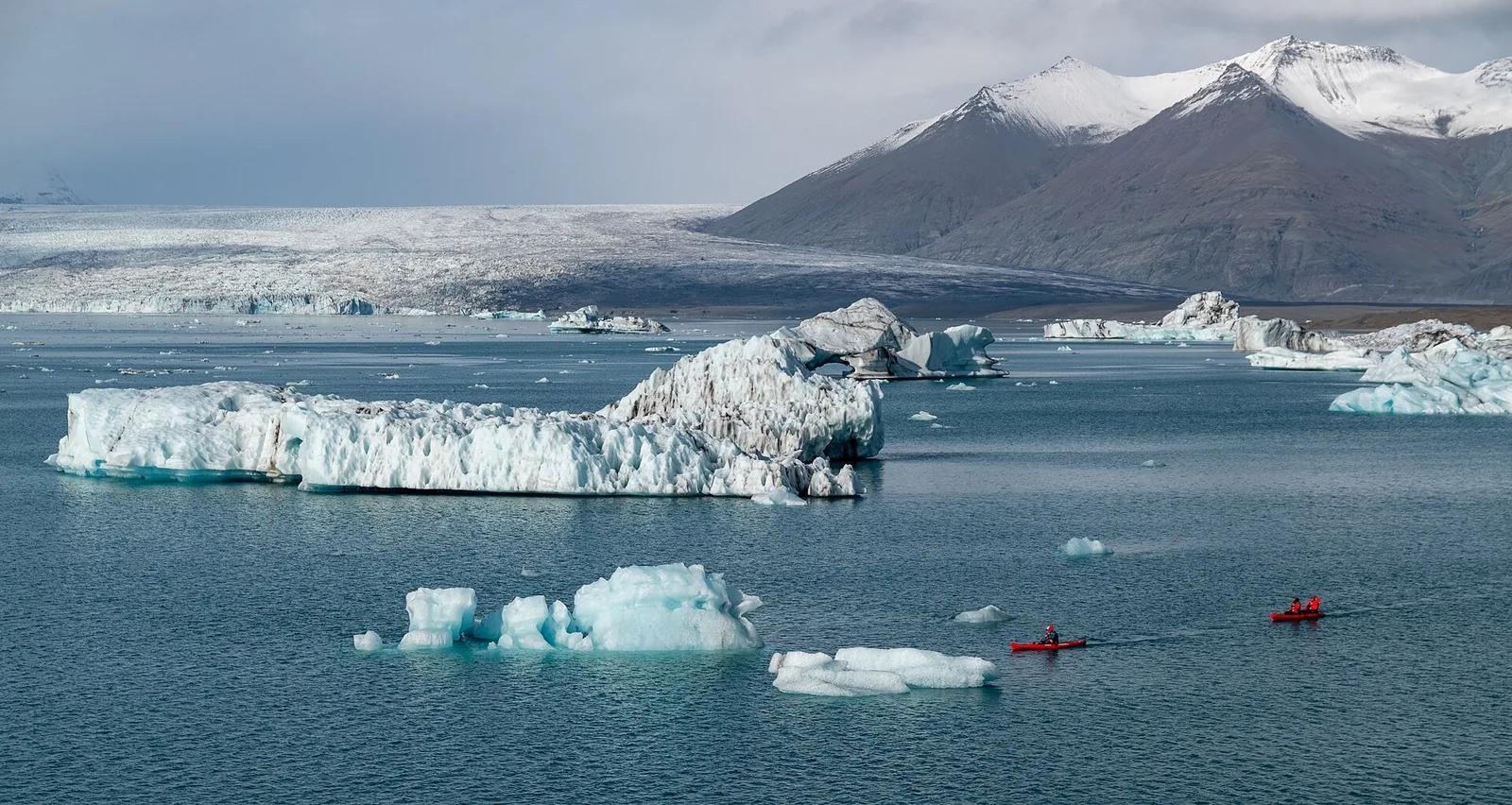 Kajaks zwischen Eisbergen auf dem Jökulsárlón im Vatnajökull-Nationalpark, dahinter die Gletscherzunge und schneebedeckte Berge