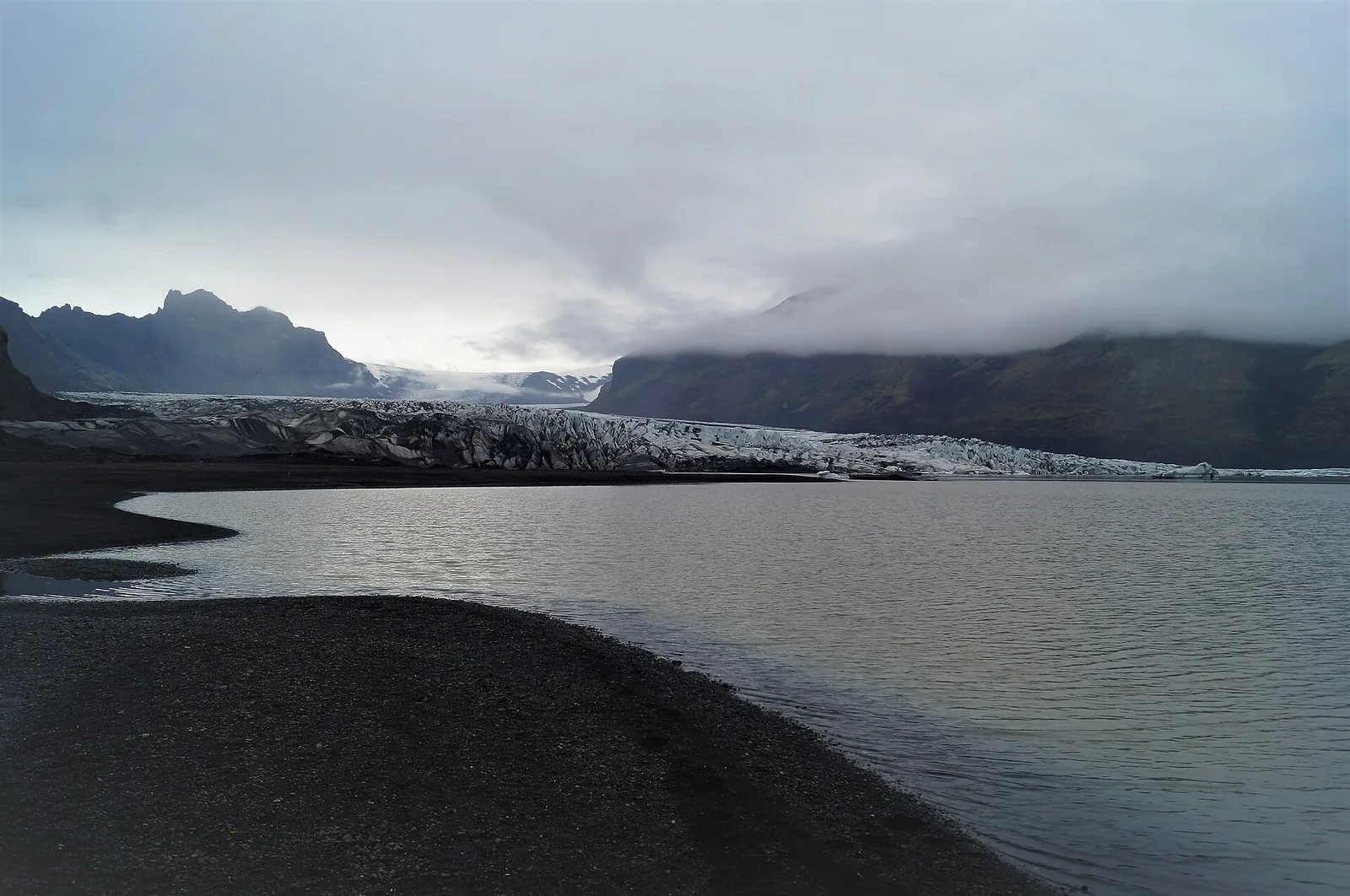 Die Zunge des Skaftafellsjökull im Vatnajökull-Nationalpark hinter einem Schmelzwassersee, dunkler Sand im Vordergrund
