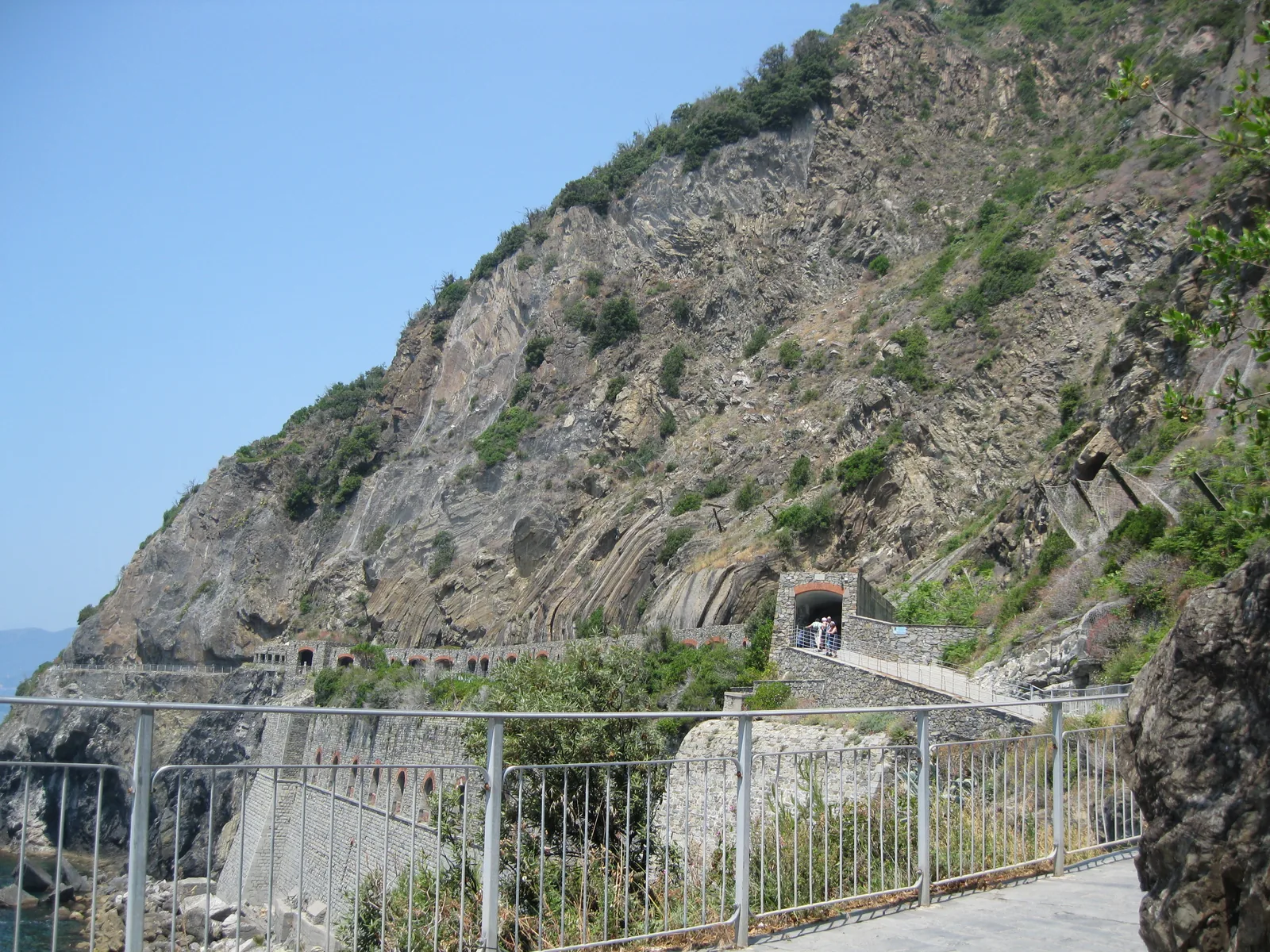 Die Via dell'Amore führt mit Geländer über eine Bahngalerie an der Felsküste bei Riomaggiore, darüber ein steiler Hang mit Macchia
