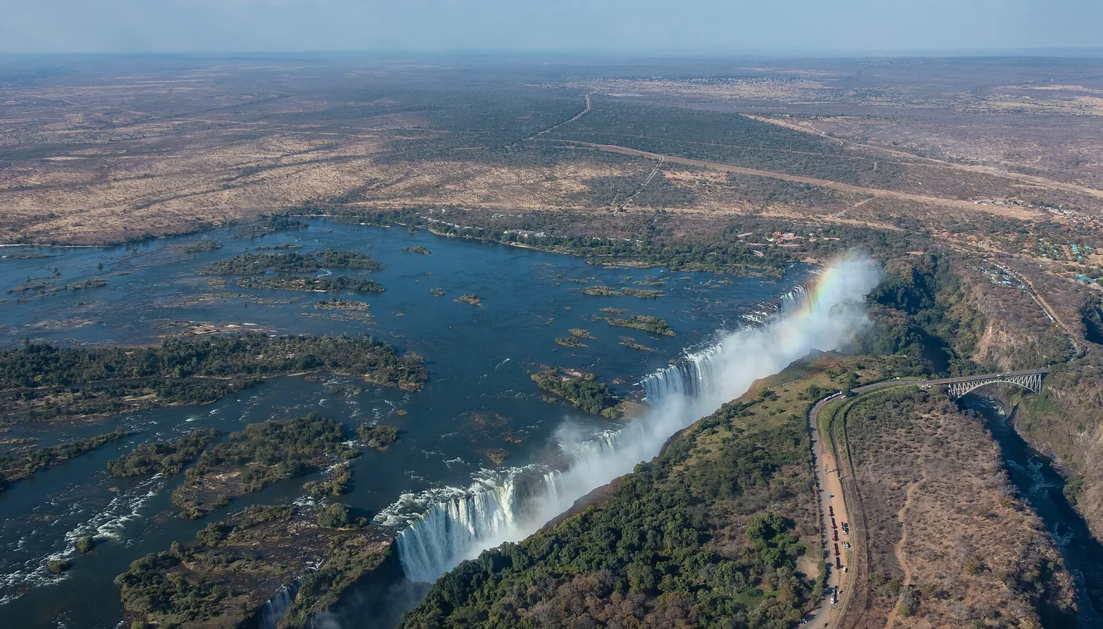 Luftbild der Victoria Falls mit dem Sambesi und seinen Inseln über der Kante, in die Schlucht stürzendes Wasser und die Straße am Ufer