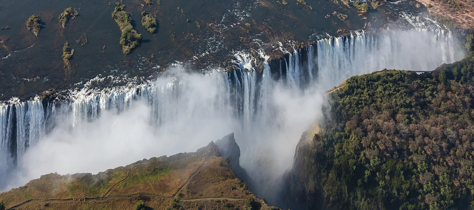Luftbild der Victoria Falls: Der Sambesi stürzt auf breiter Front in die erste Schlucht, Gischt steigt auf, rechts der grüne Regenwald auf der simbabwischen Seite
