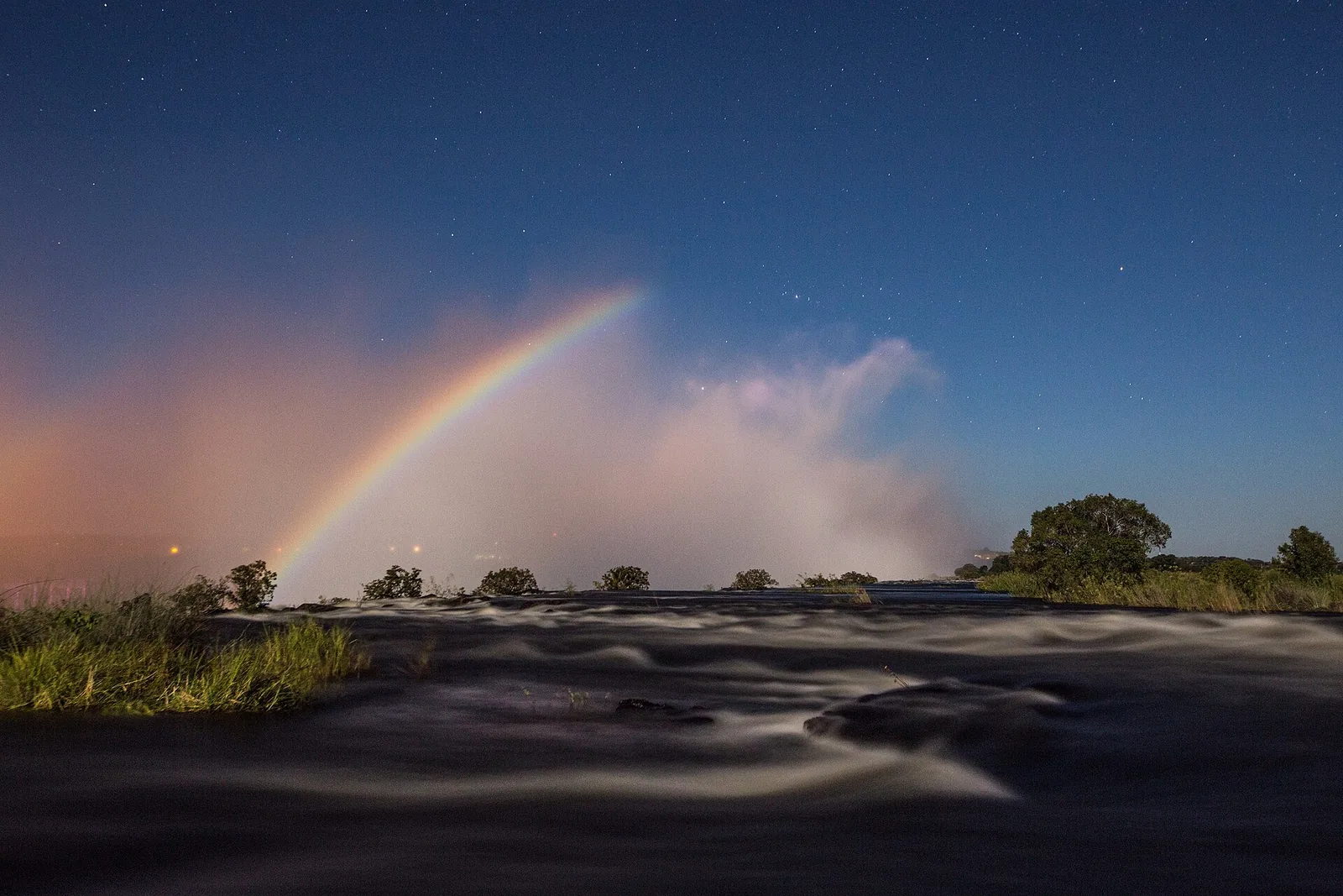 Ein Mondregenbogen spannt sich nachts über die Gischt der Victoria Falls, darunter der dunkle Sambesi und Sterne am Himmel
