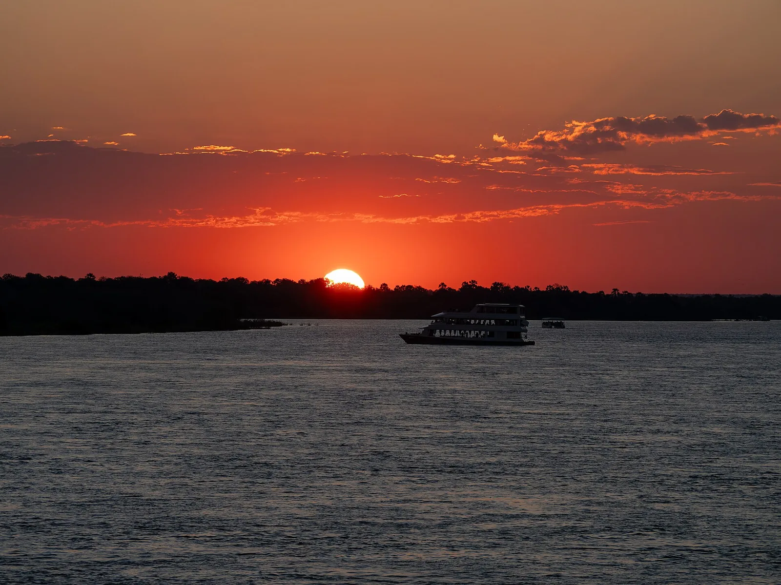 Sonnenuntergang auf dem Sambesi oberhalb der Victoria Falls, ein Ausflugsboot vor dem dunklen Ufer und rotem Himmel