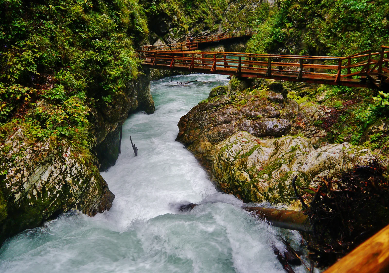 Wildwasser zwischen hellen Felsblöcken, oben ein Steg entlang der Wand