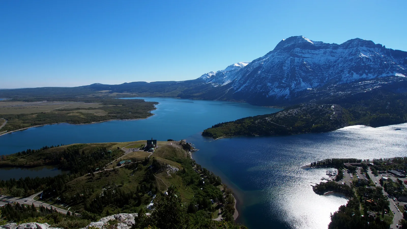 Blick vom Bear's Hump über den Upper Waterton Lake: links die flache Prärie, rechts die schneegestreifte Bergwand, auf der Anhöhe das Prince of Wales Hotel