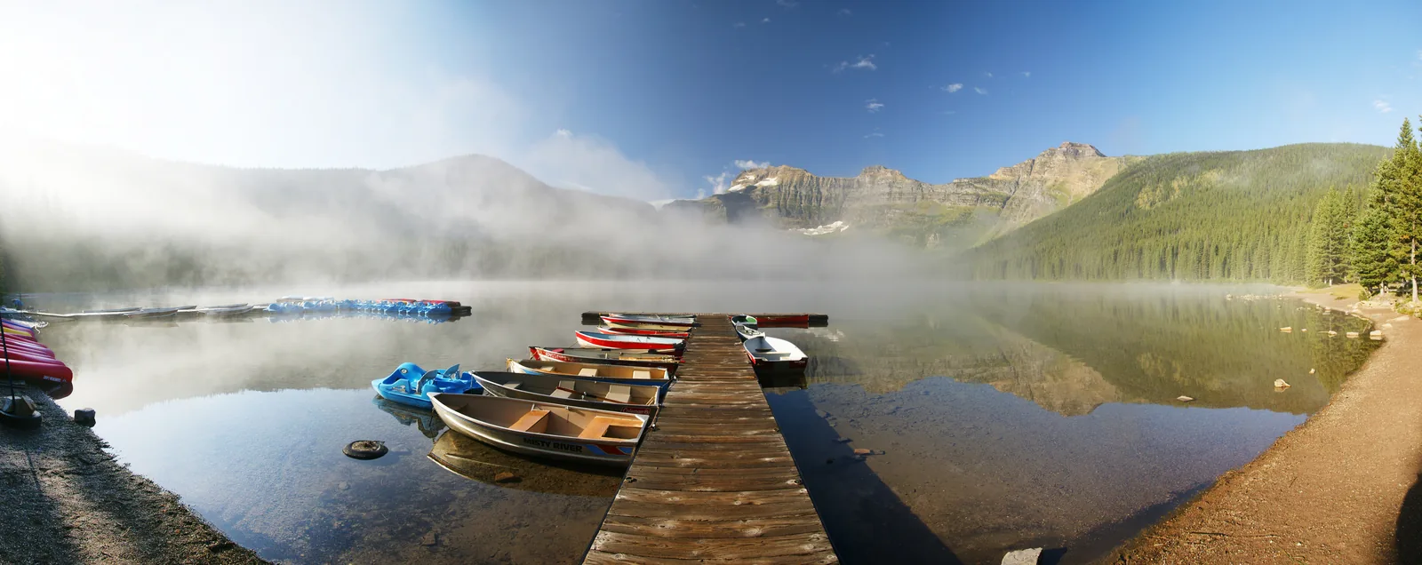 Früher Morgen am Cameron Lake: Nebel über dem Wasser, ein Steg mit aufgereihten Kanus, dahinter bewaldete Berghänge