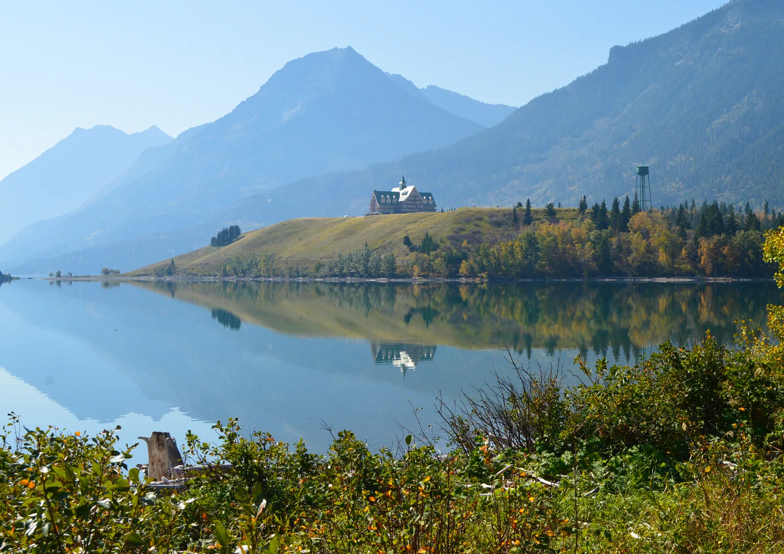 Das Prince of Wales Hotel auf einer Anhöhe über dem spiegelglatten Waterton-See, dahinter die Berge, im Vordergrund herbstliches Ufergebüsch