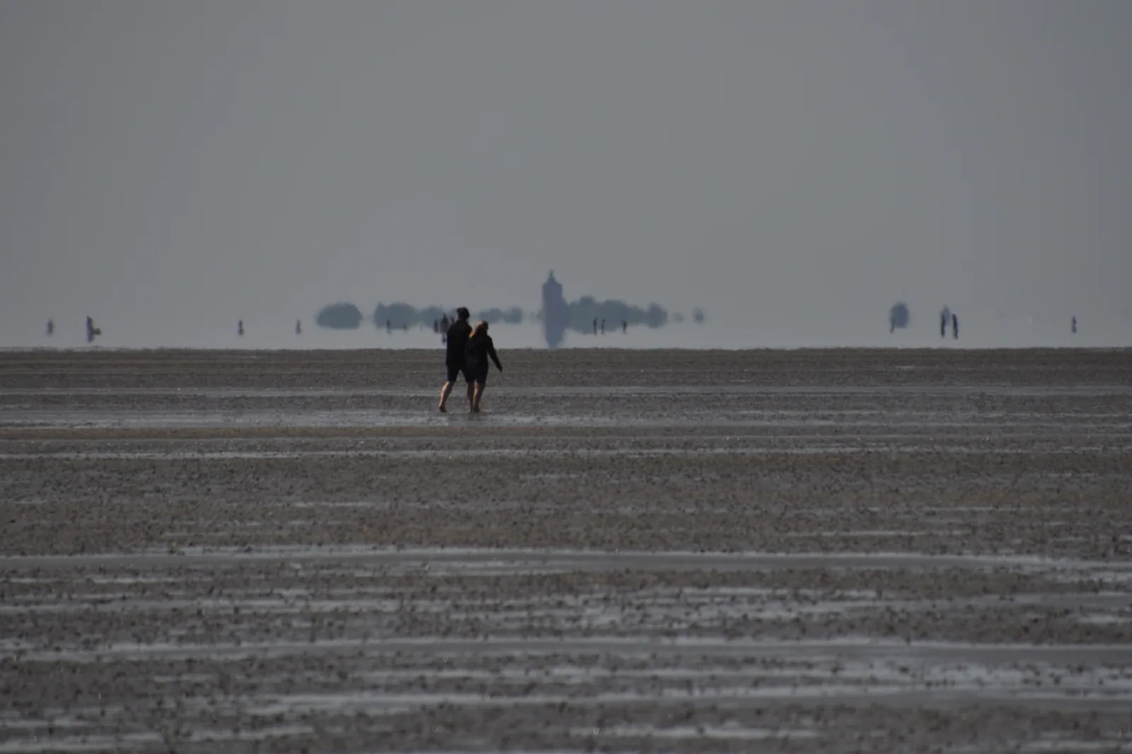 Zwei Menschen bei einer Wattwanderung nach Neuwerk, am Horizont spiegeln sich Turm und Insel im Dunst über dem Watt