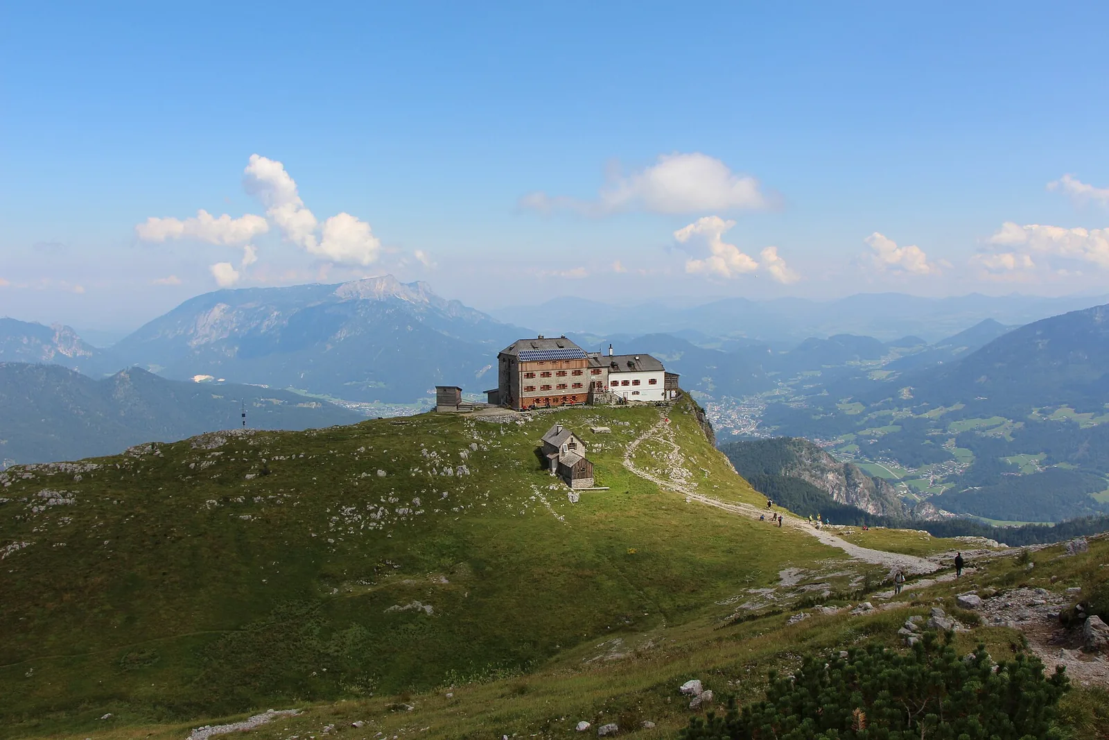 Das Watzmannhaus auf einem grünen Bergrücken im Nationalpark Berchtesgaden, dahinter das Berchtesgadener Tal und ferne Gipfel