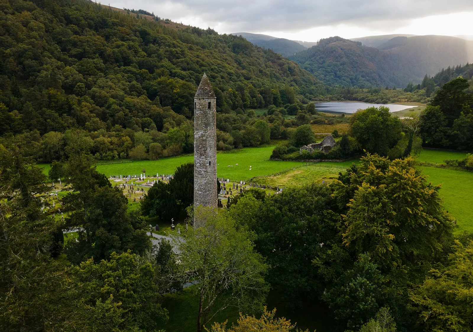 Luftbild der Klostersiedlung Glendalough im Wicklow-Mountains-Nationalpark mit dem Rundturm zwischen Bäumen und Gräbern