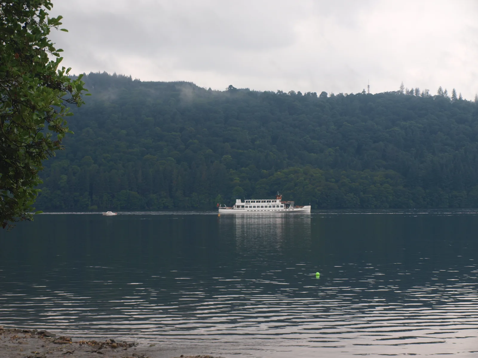 Ein Ausflugsschiff auf dem Windermere vor dem bewaldeten Westufer, gesehen vom Ufer in Bowness-on-Windermere