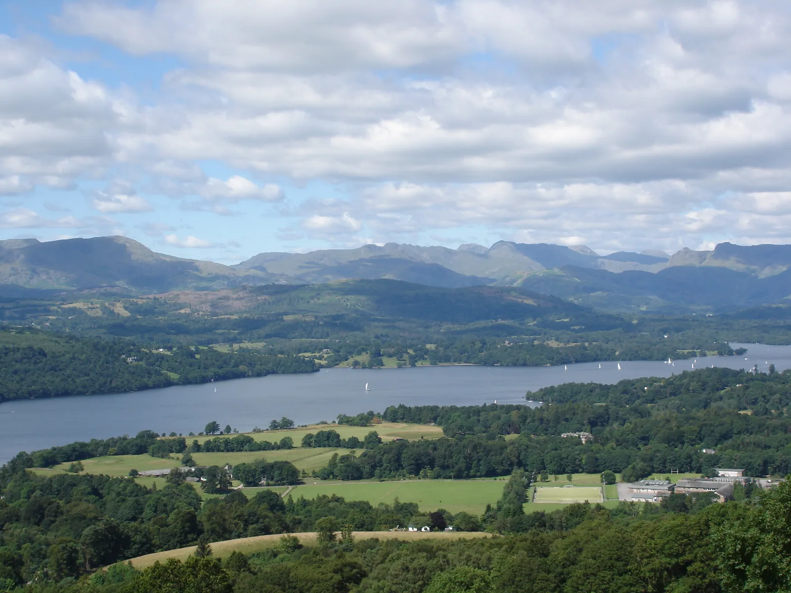 Blick von einem Hügel über den Windermere im Lake-District-Nationalpark, dahinter Wiesen, Wälder und die Fells
