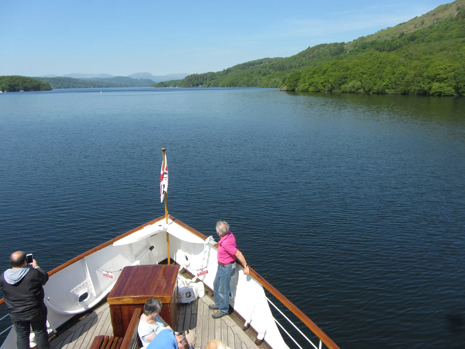 Fahrgäste am Bug eines Ausflugsschiffs auf dem Windermere im Lake District, vor ihnen der See und bewaldete Ufer