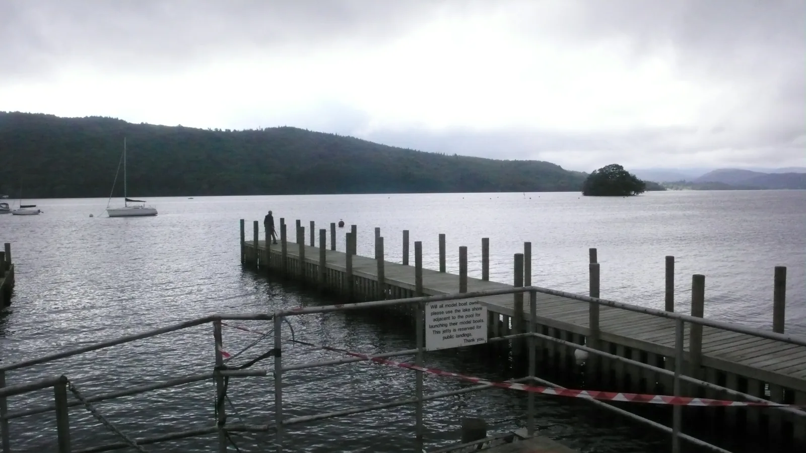 Ein Holzsteg ragt in den Windermere im Lake District, dahinter eine kleine Insel und Hügel unter Wolken