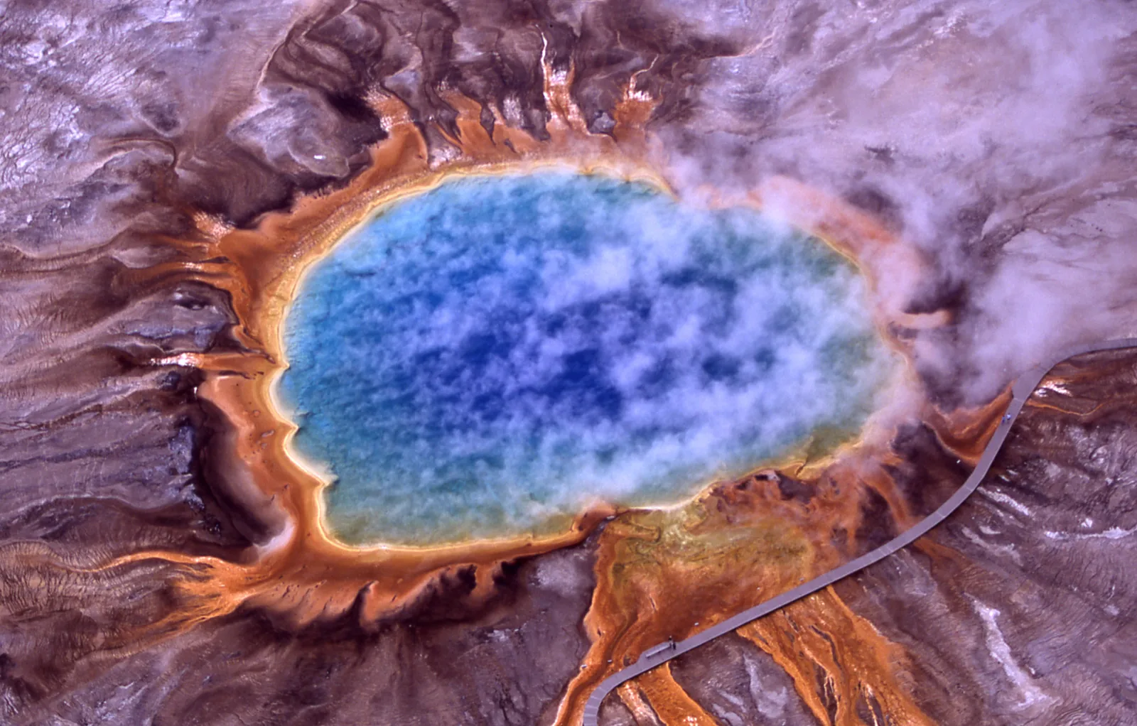 Luftaufnahme einer kreisrunden heißen Quelle mit blauem Zentrum und orangegelben Rändern inmitten heller Sinterflächen