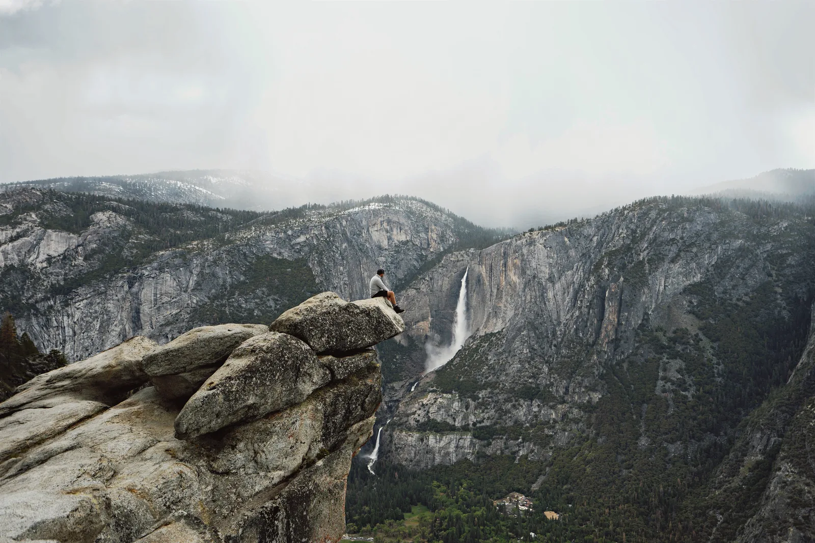 Ein Mensch sitzt auf einem vorspringenden Felsen am Glacier Point, gegenüber stürzt der Yosemite Fall über die Talwand