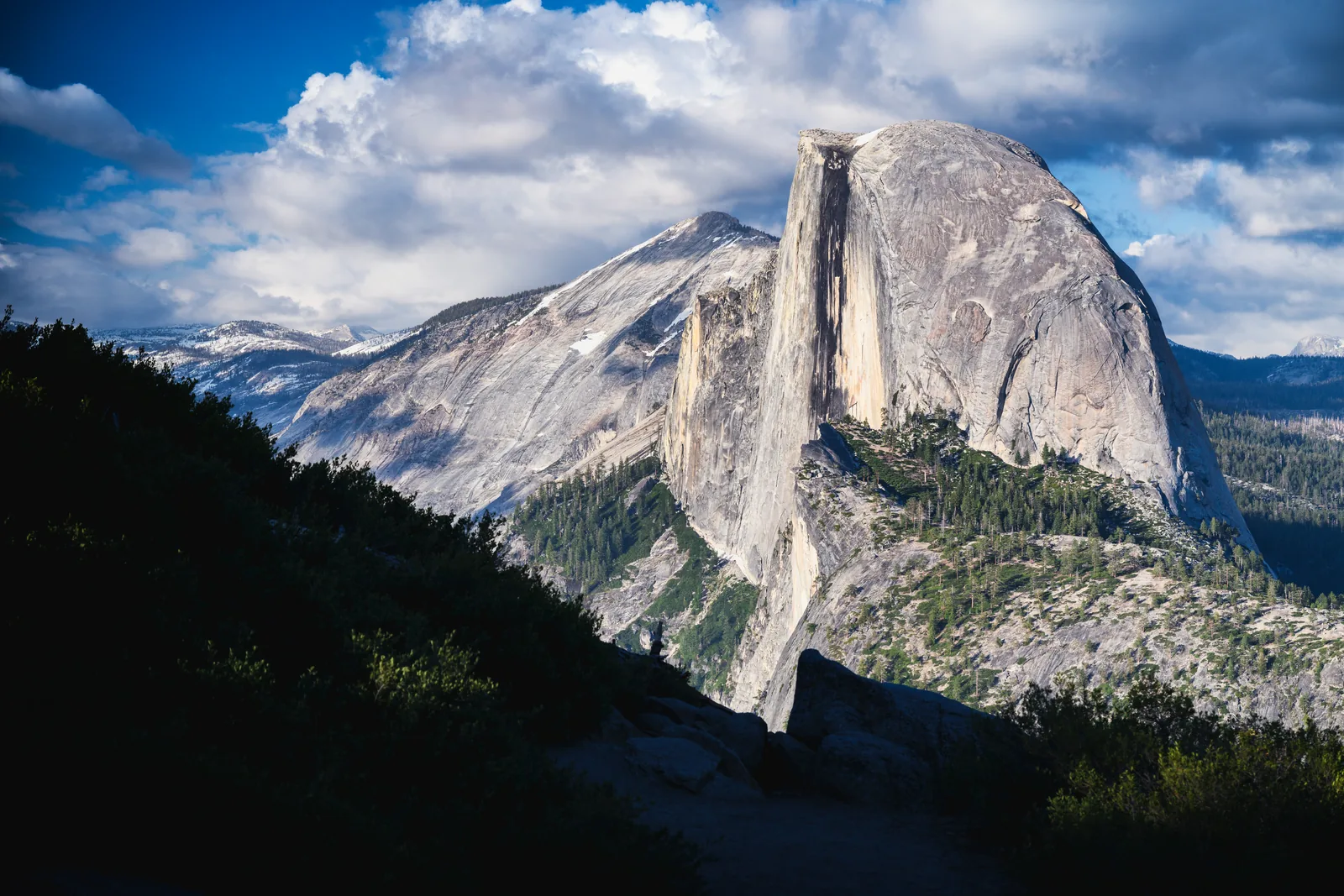 Der Half Dome im Abendlicht: eine senkrecht abgebrochene Granitkuppe über bewaldeten Hängen, dahinter schneefleckige Berge