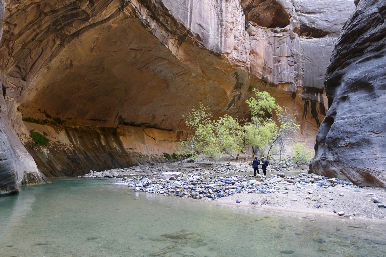 Türkisfarbenes Wasser zwischen hohen, glatt geschliffenen Sandsteinwänden, am Ufer zwei Wanderer und ein einzelner Baum